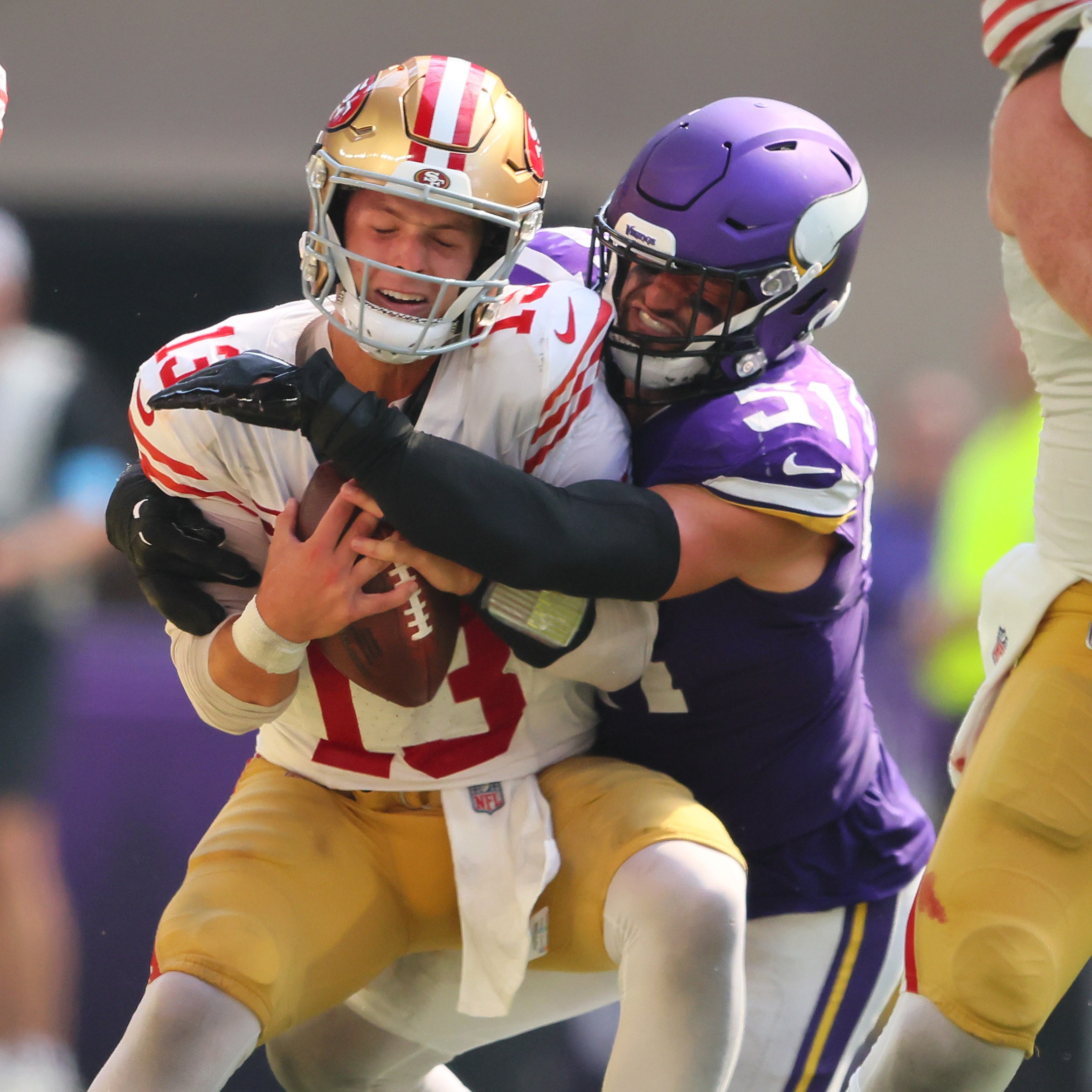 A football player in a purple uniform tackles a player in a white and red uniform holding a football. The scene is intense, with other players around them.