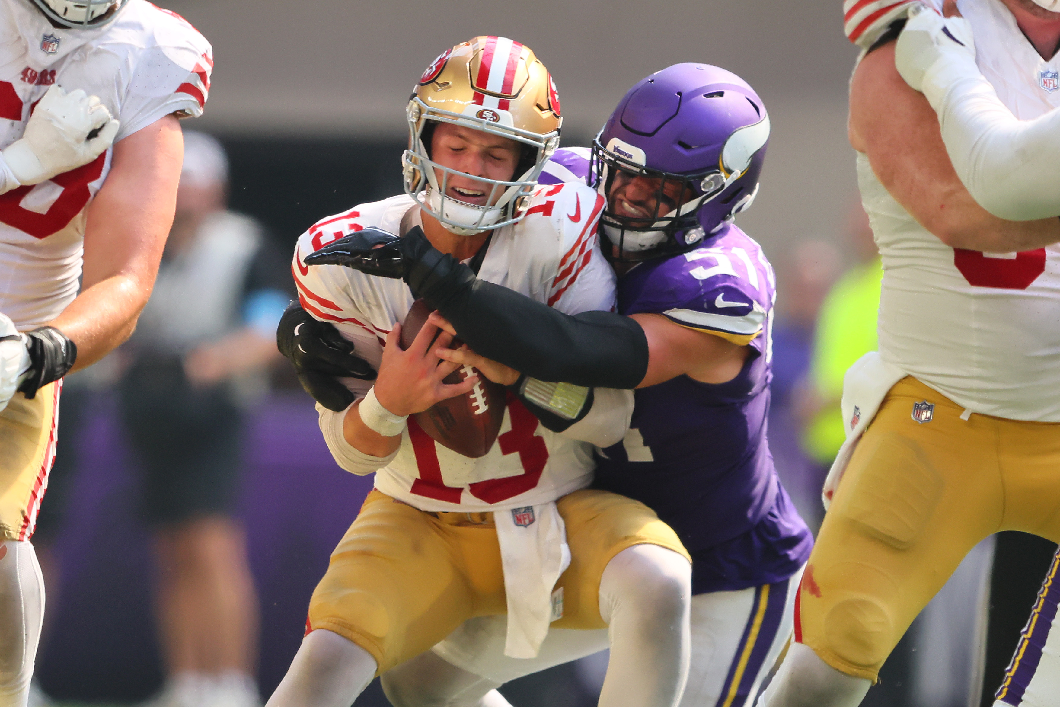 A football player in a purple uniform tackles a player in a white and red uniform holding a football. The scene is intense, with other players around them.