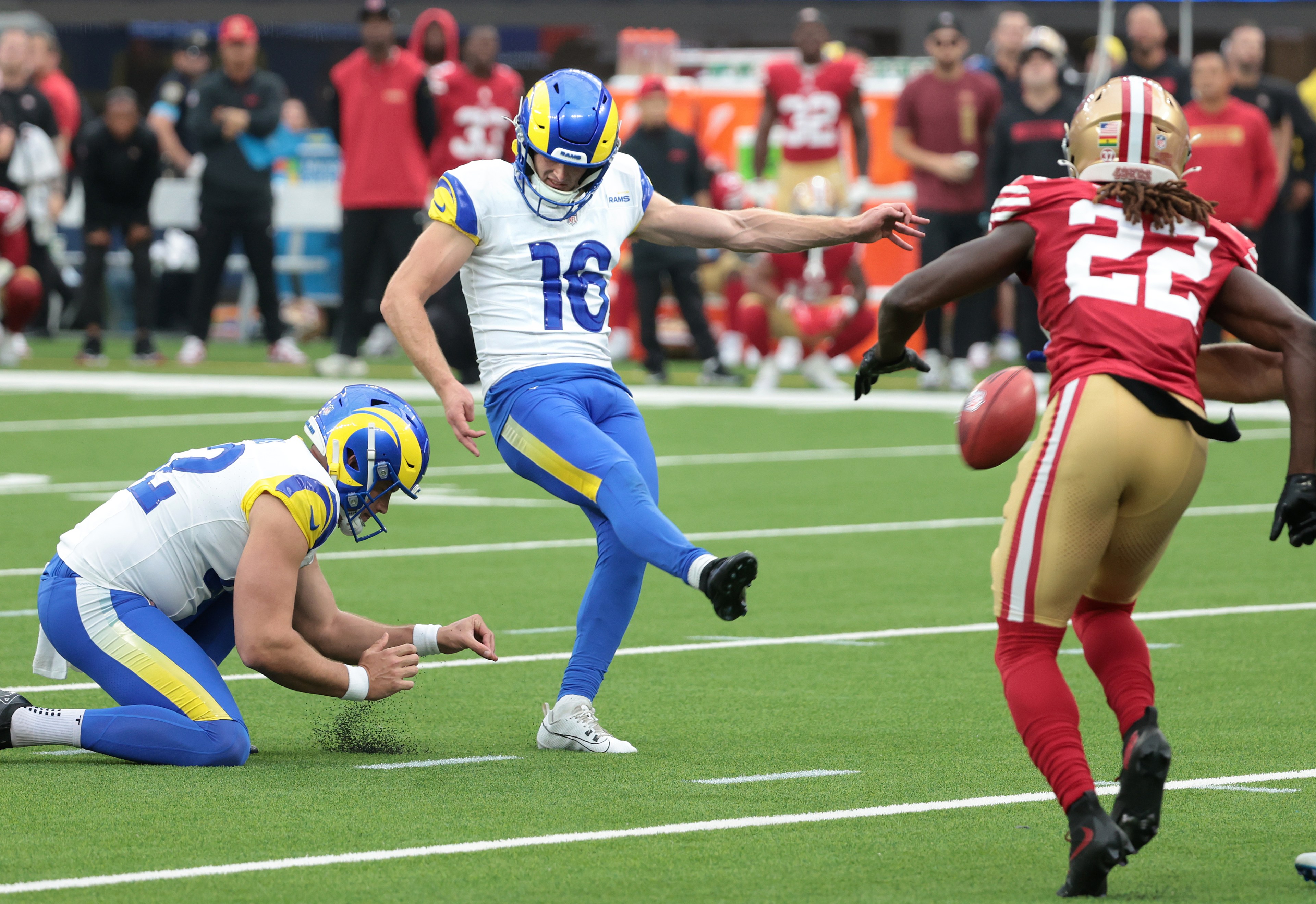 A football kicker in blue and white is about to kick the ball held by a teammate as a player in red and gold rushes towards them on a green field.