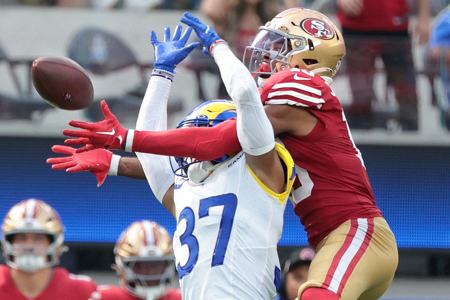 Two American football players leap to catch a football mid-air; one wears a red and gold uniform, and the other wears a blue and white uniform.