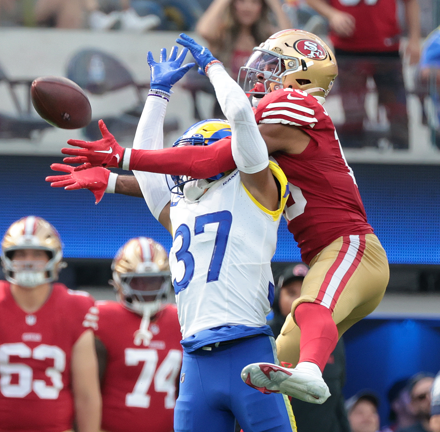 Two American football players leap to catch a football mid-air; one wears a red and gold uniform, and the other wears a blue and white uniform.