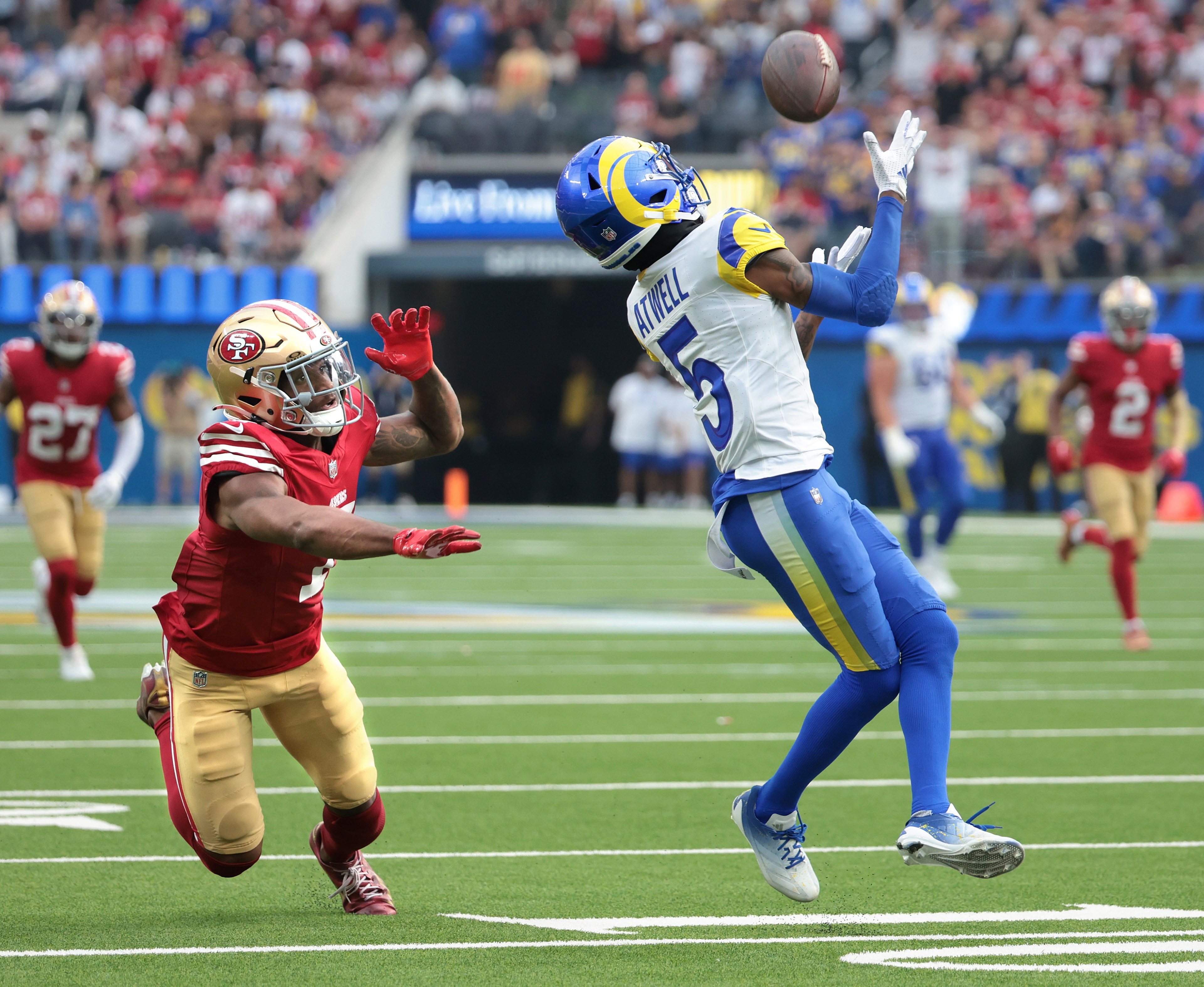 A football player in a white and blue uniform is reaching to catch the ball, while another player in a red uniform attempts to block him on the field.