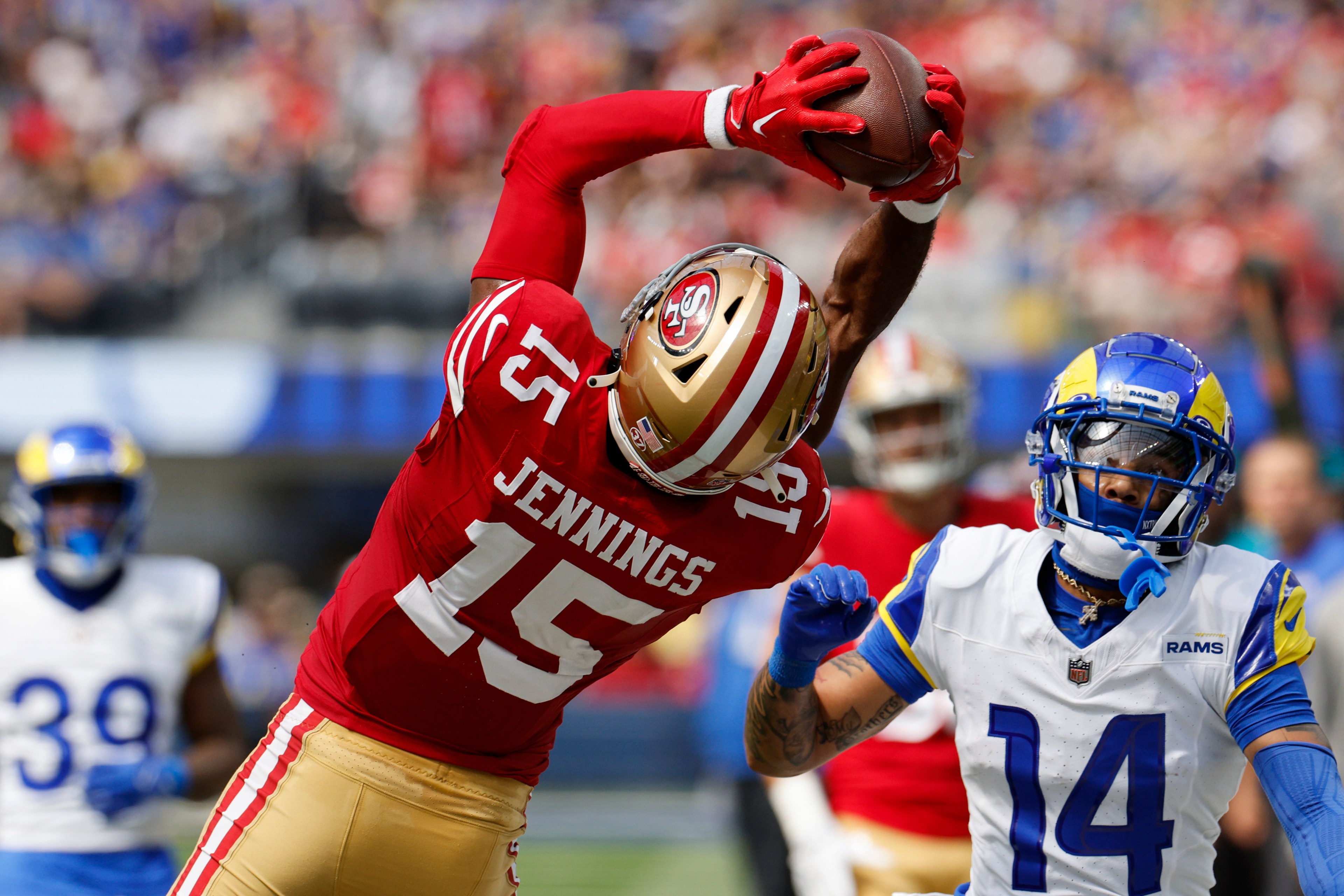 A football player in a red and gold uniform labeled &quot;Jennings 15&quot; leaps to catch the ball while a player in a white and blue uniform attempts to defend.