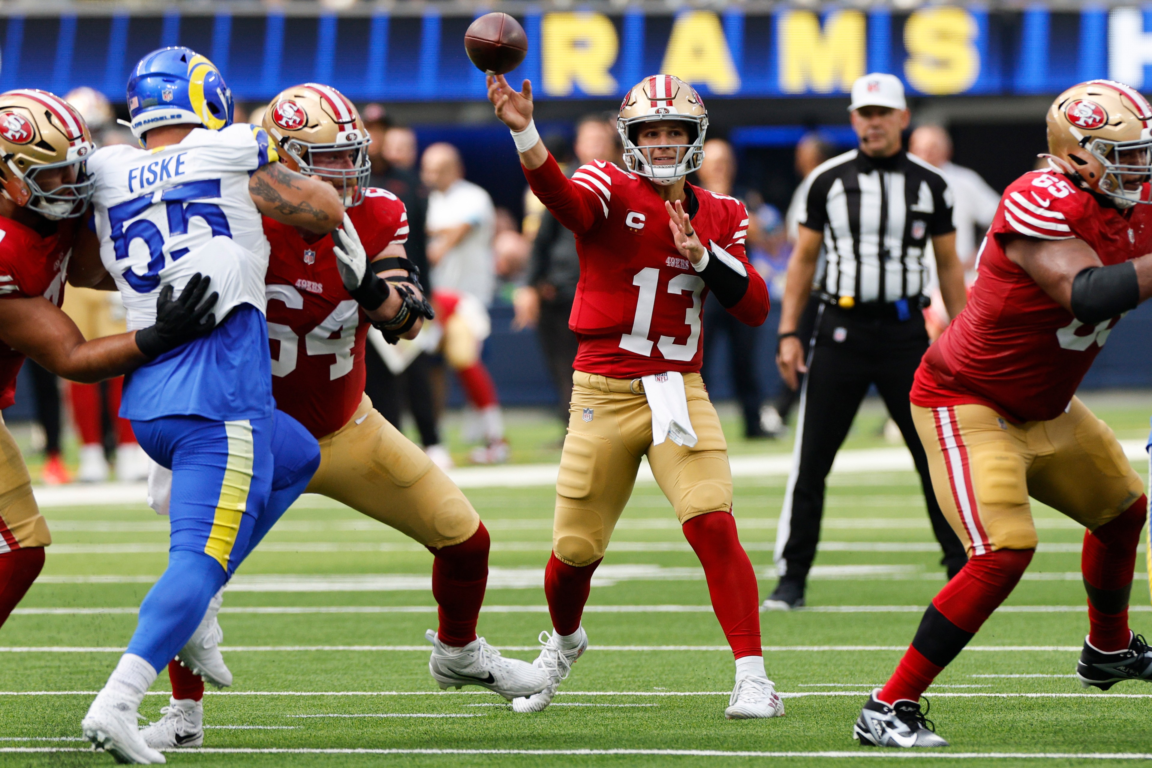 A football player in a red uniform throws a pass while teammates block an opponent in a blue and white uniform; a referee stands in the background.