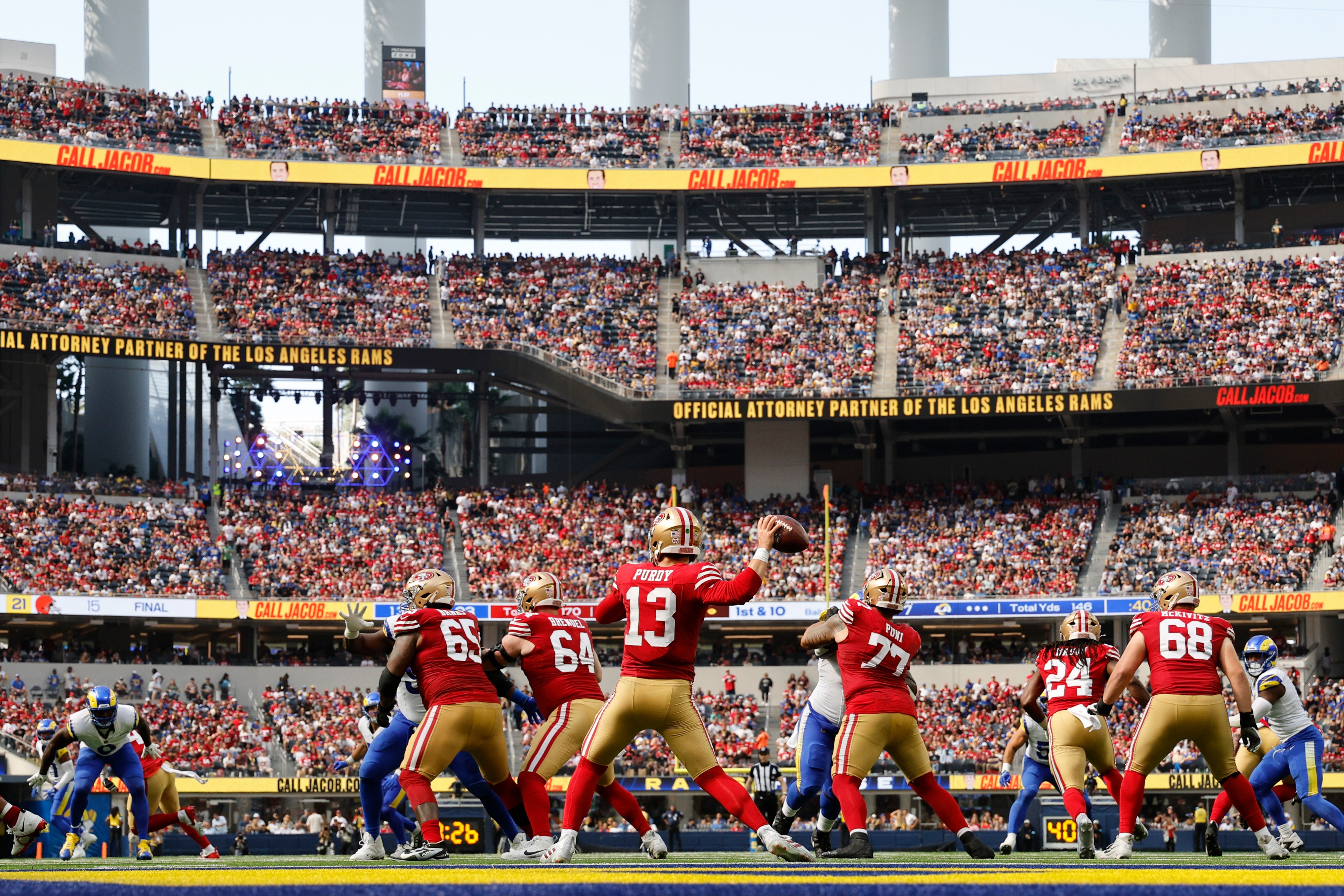 A football game is in action with a San Francisco 49ers quarterback preparing to throw the ball. Teammates block opposing Los Angeles Rams players in a crowded stadium.