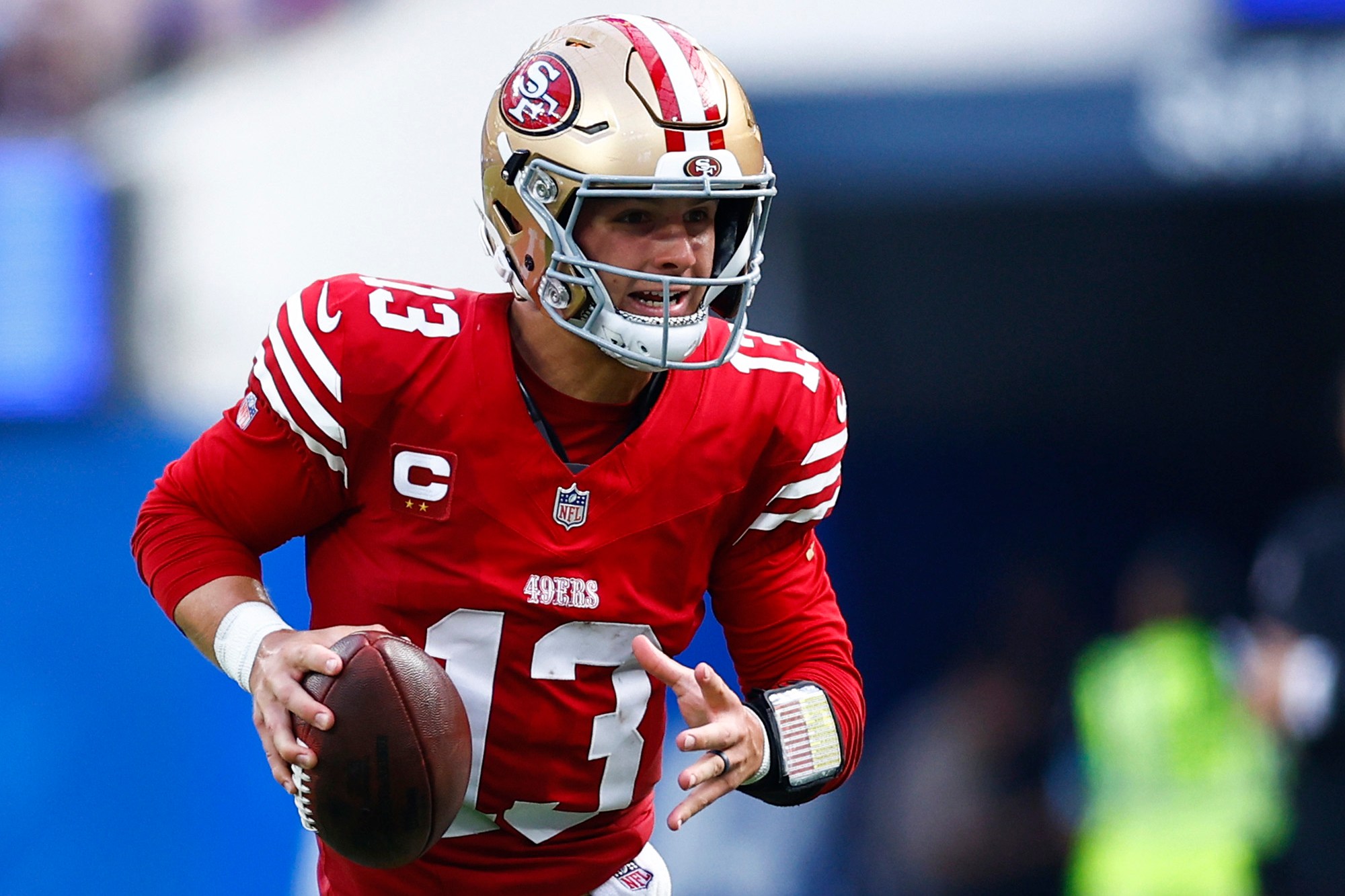 A football player in a red and gold uniform is running with the ball, looking ahead, likely to pass. The field is green turf, and the background is blurred.