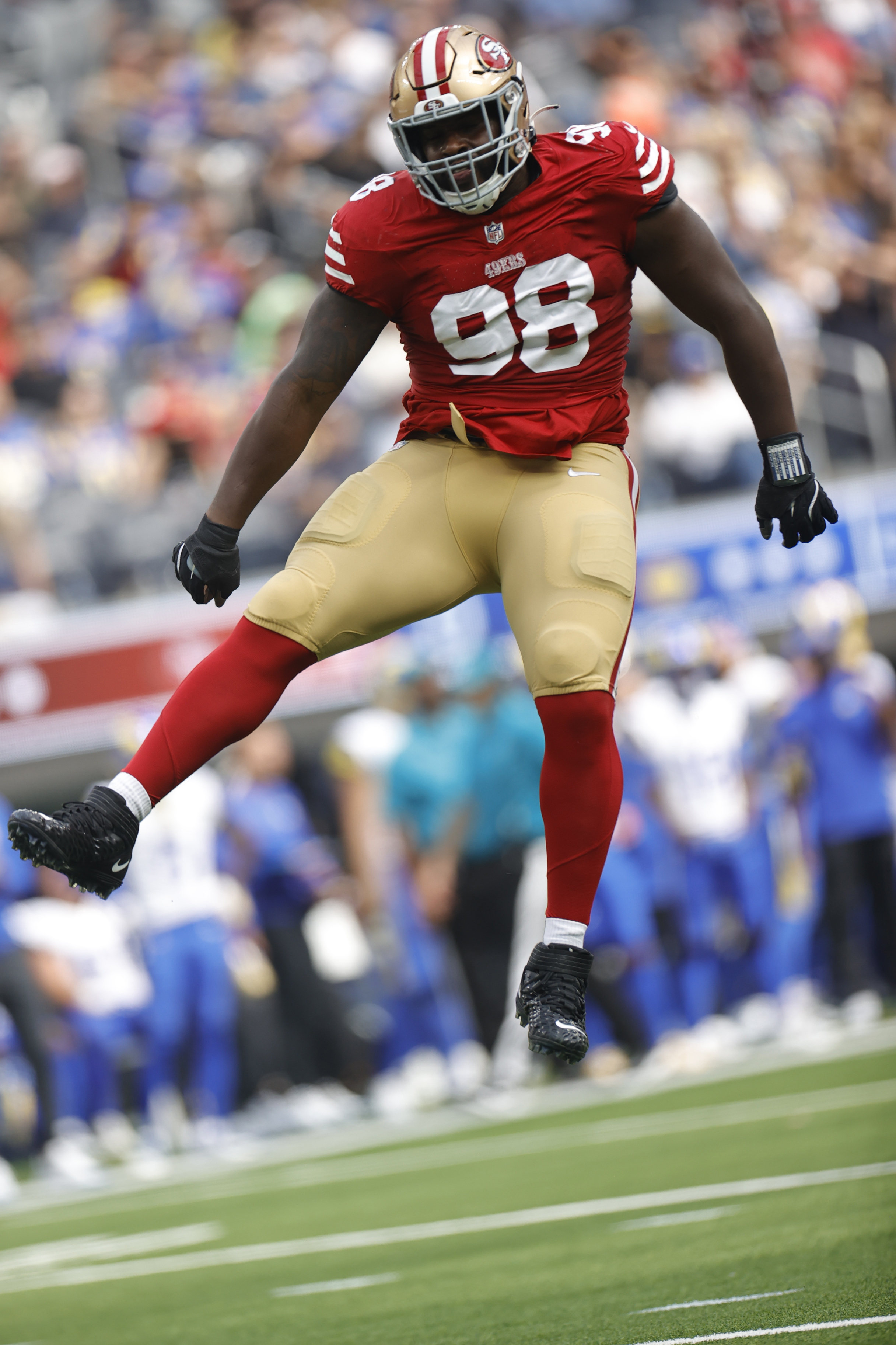 A football player in a red and gold uniform is jumping in the air on a field during a game. The background is blurred, showing fans and other players.