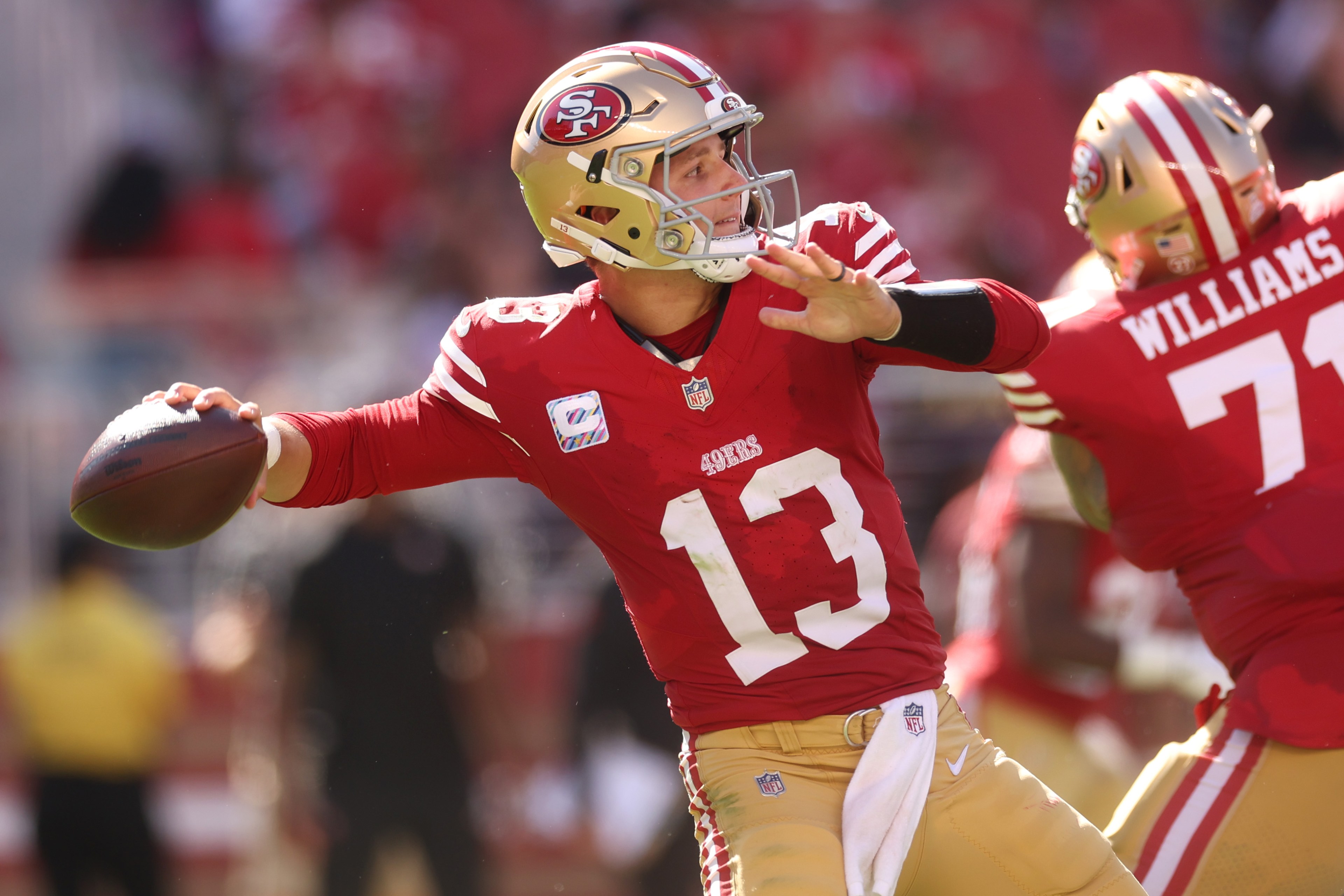 A football player in a red jersey, numbered 13, prepares to throw the ball during a game, with another player, numbered 71, blocking in the background.