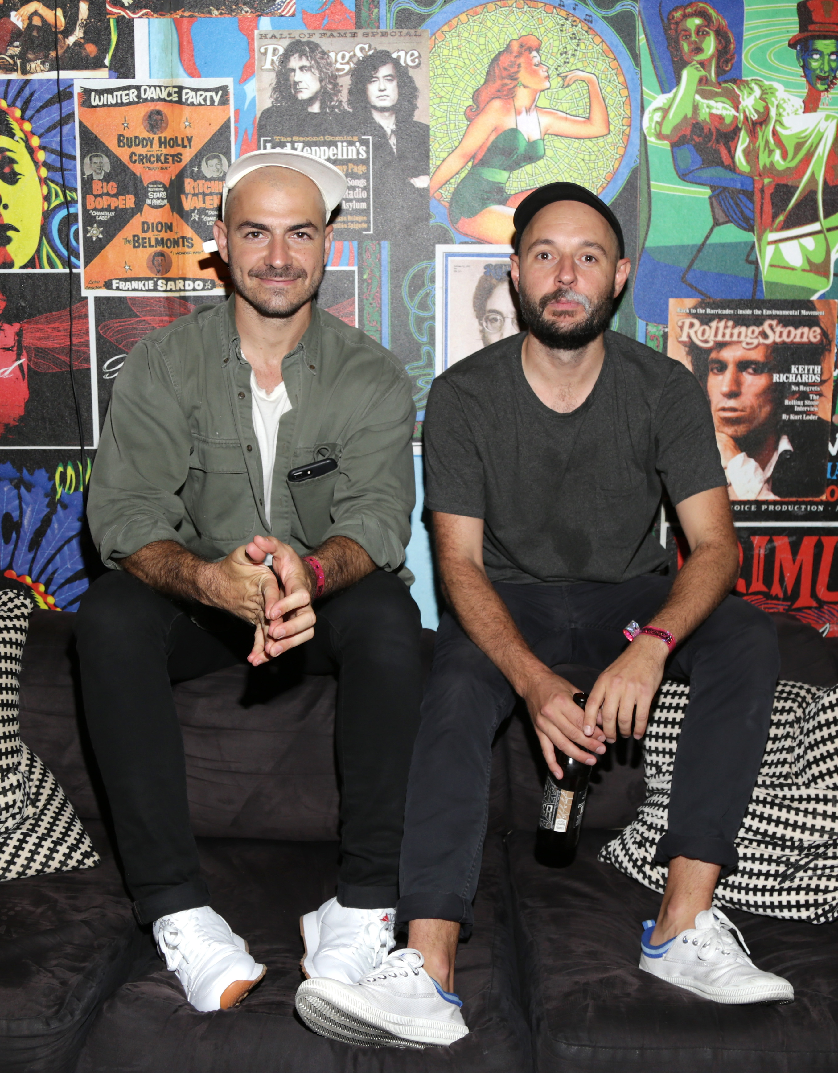 Two men sit on a black couch in front of a colorful wall covered with various posters. One wears a green shirt and white sneakers, the other a gray shirt holding a bottle.