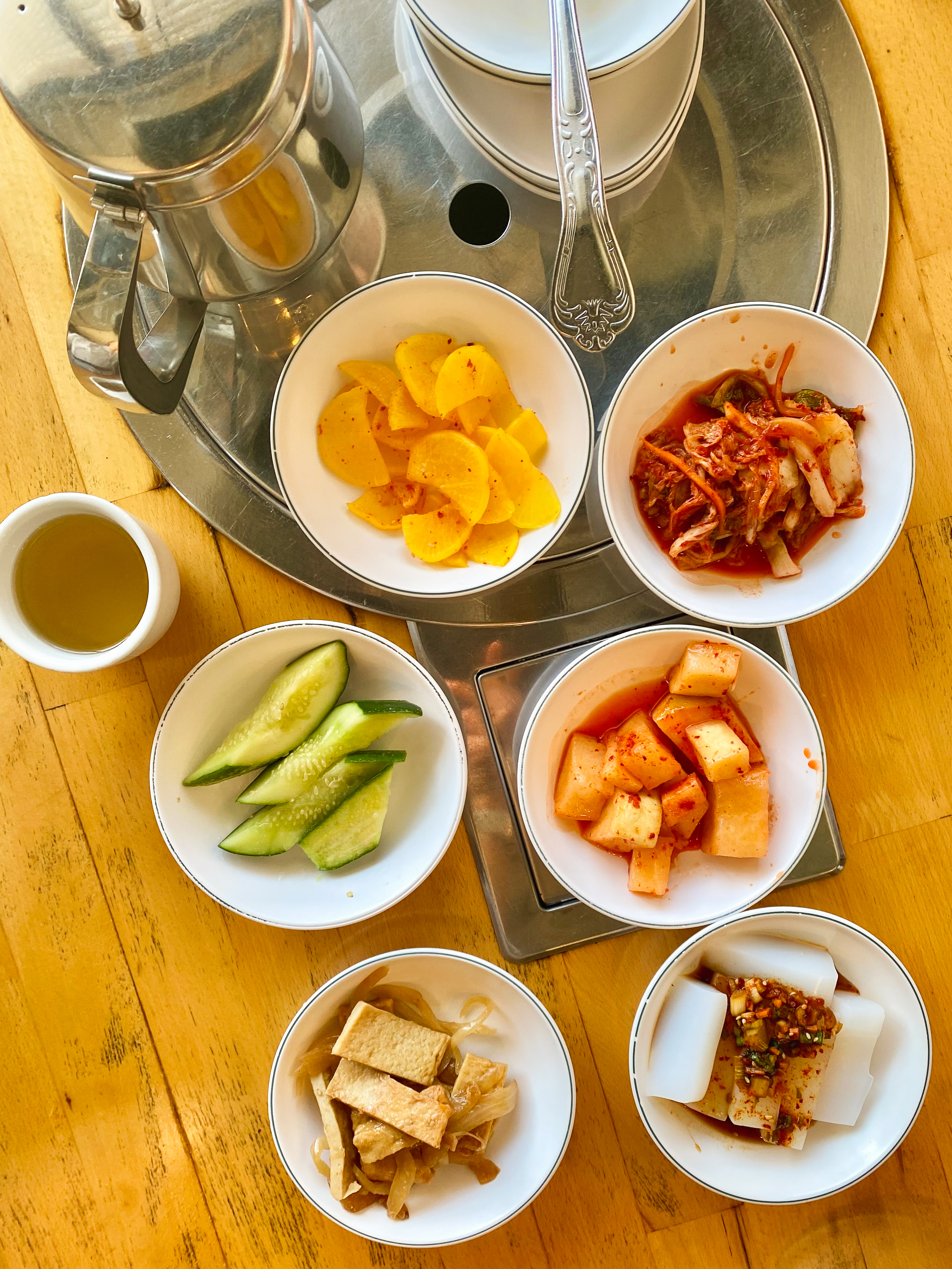 This image shows a wooden table set with six small bowls containing various side dishes, a metal teapot, a tea cup, and stacked dishes with chopsticks.