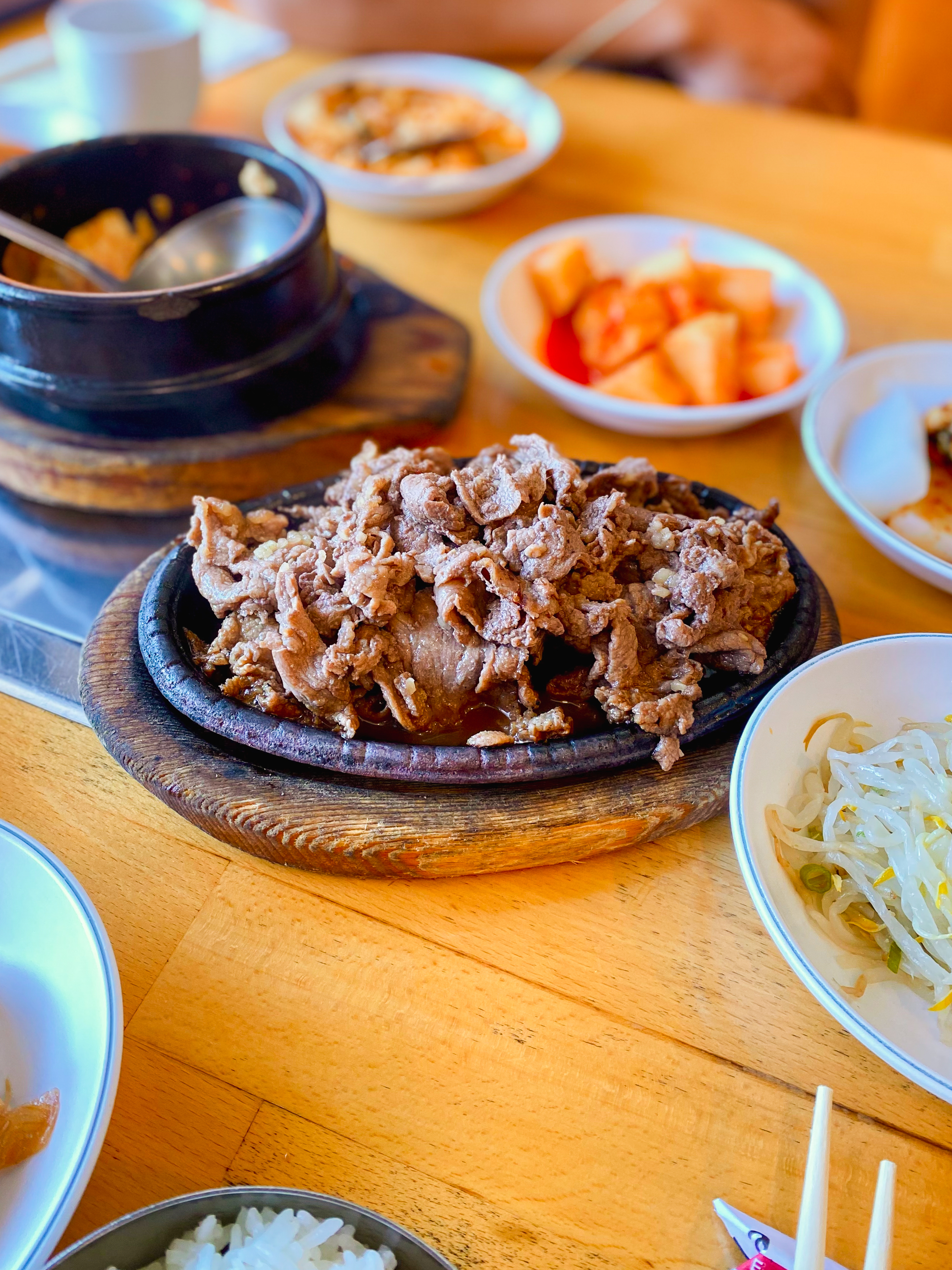 A sizzling platter of cooked beef is surrounded by various side dishes, including kimchi, bean sprouts, and rice on a wooden table.