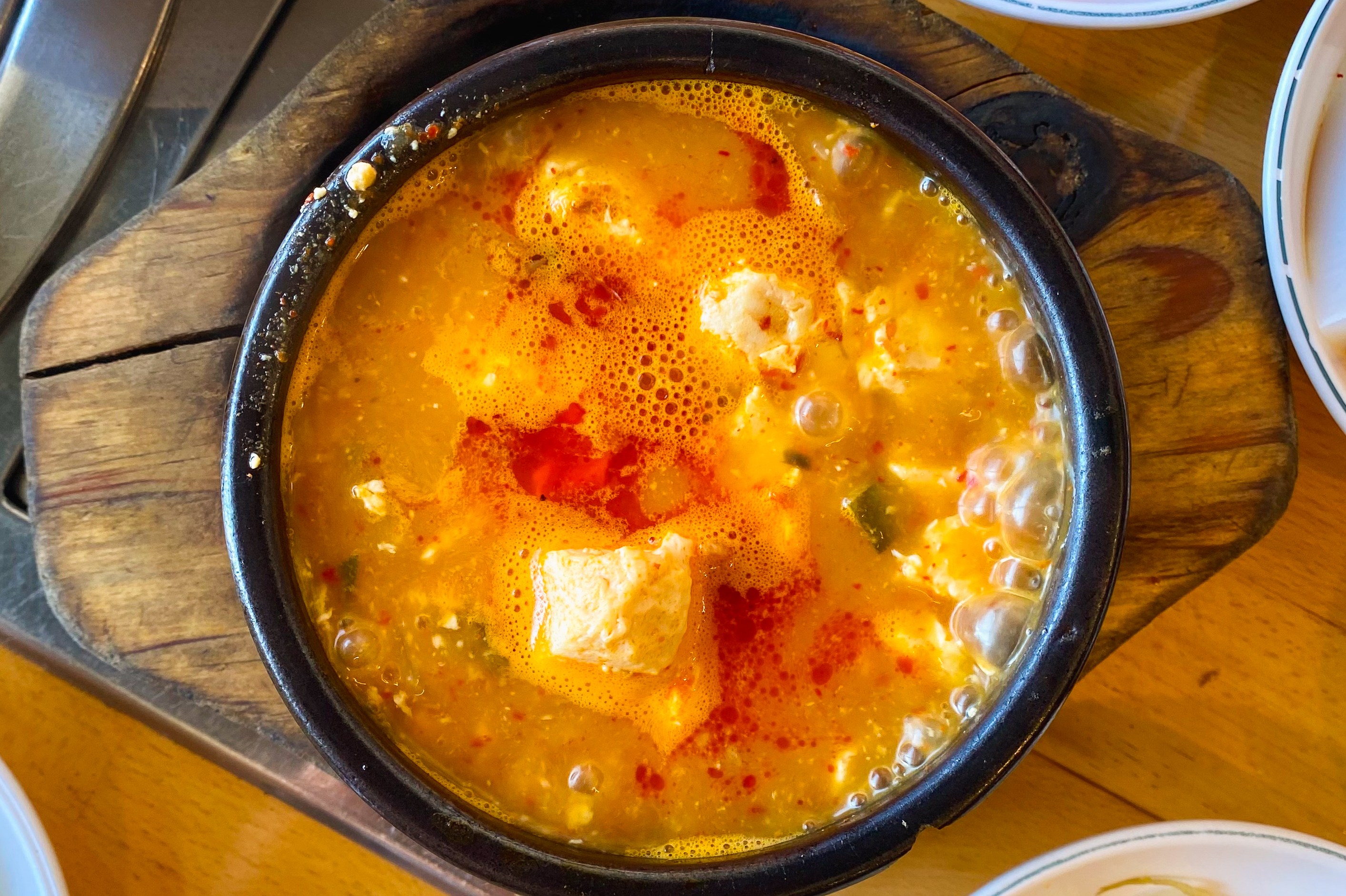 A bubbling pot of stew is surrounded by small bowls containing side dishes like kimchi, radish cubes in red sauce, and bean sprouts on a wooden table.