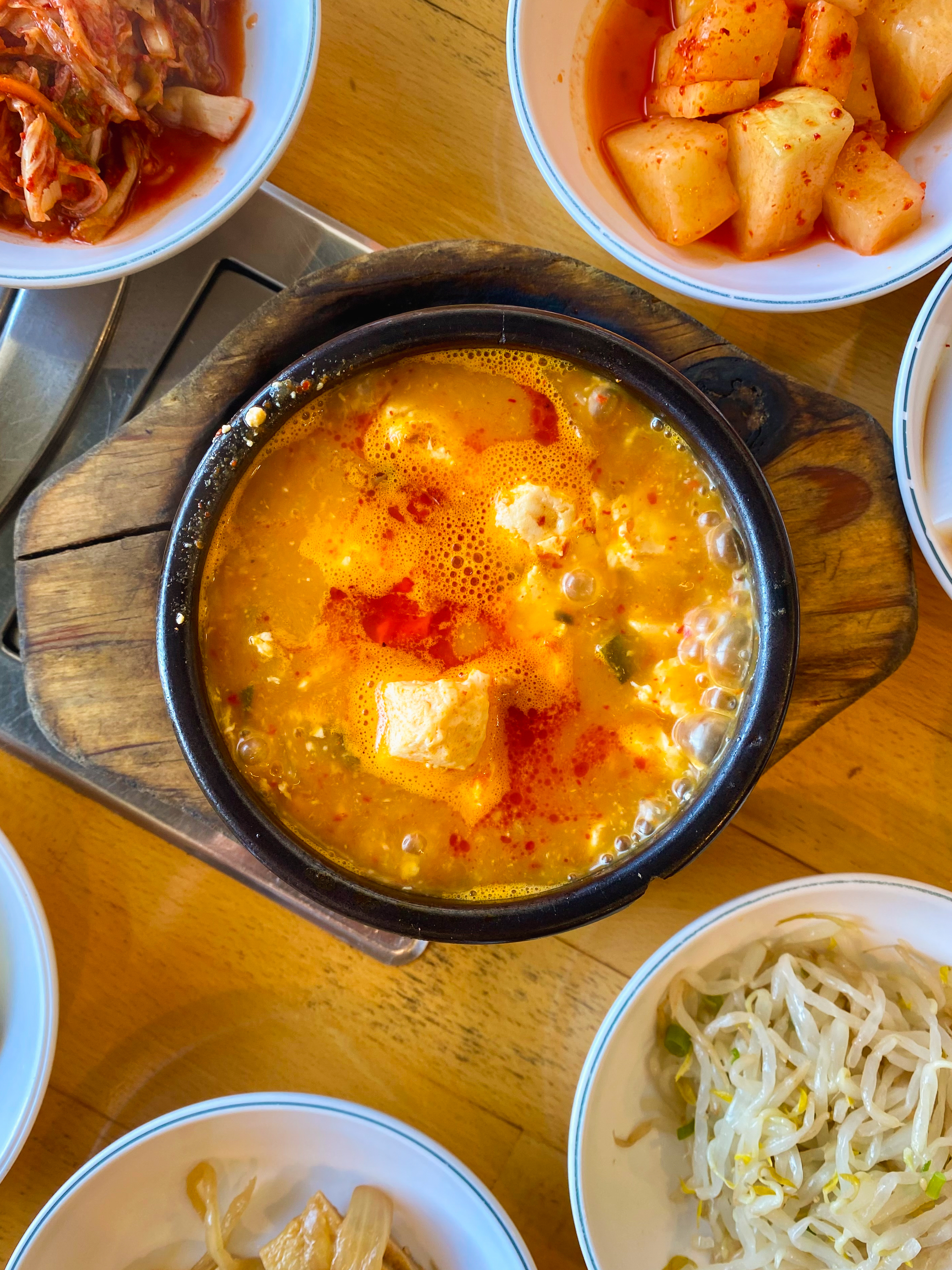 A bubbling pot of stew is surrounded by small bowls containing side dishes like kimchi, radish cubes in red sauce, and bean sprouts on a wooden table.