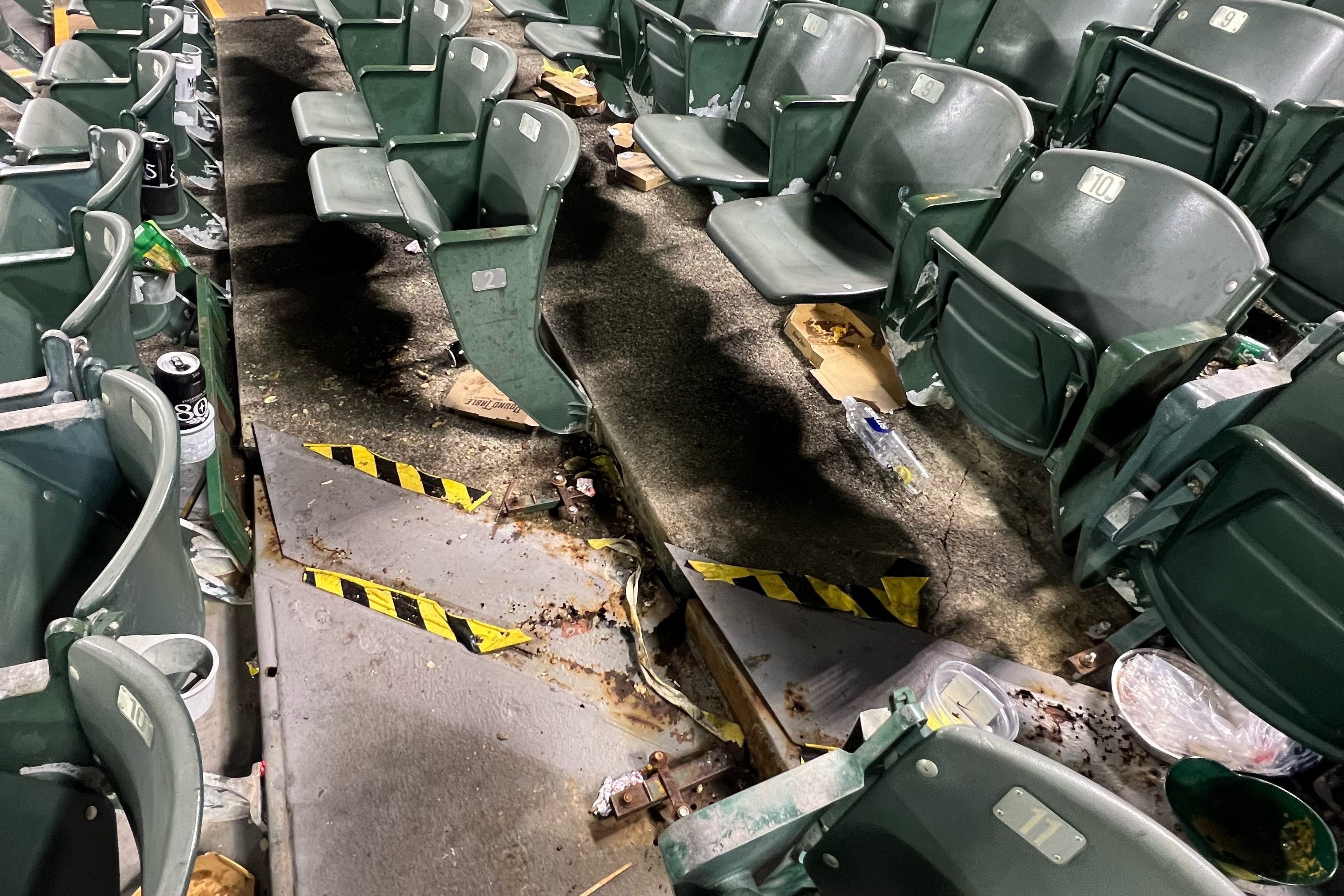 A section of green stadium seats is shown, with a fire-damaged area in the middle. The seats are surrounded by trash, including food containers and cans.