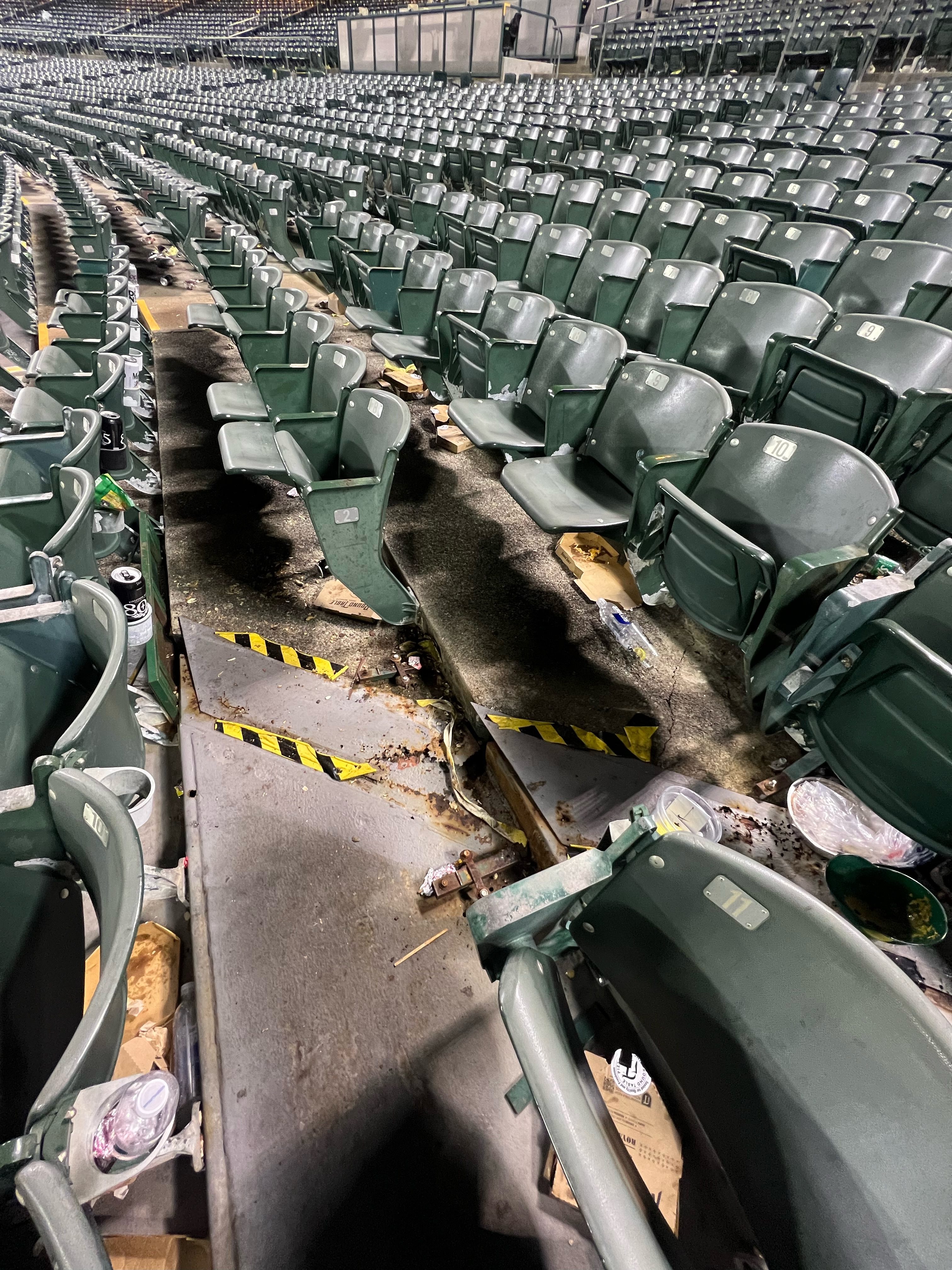 A section of green stadium seats is shown, with a fire-damaged area in the middle. The seats are surrounded by trash, including food containers and cans.