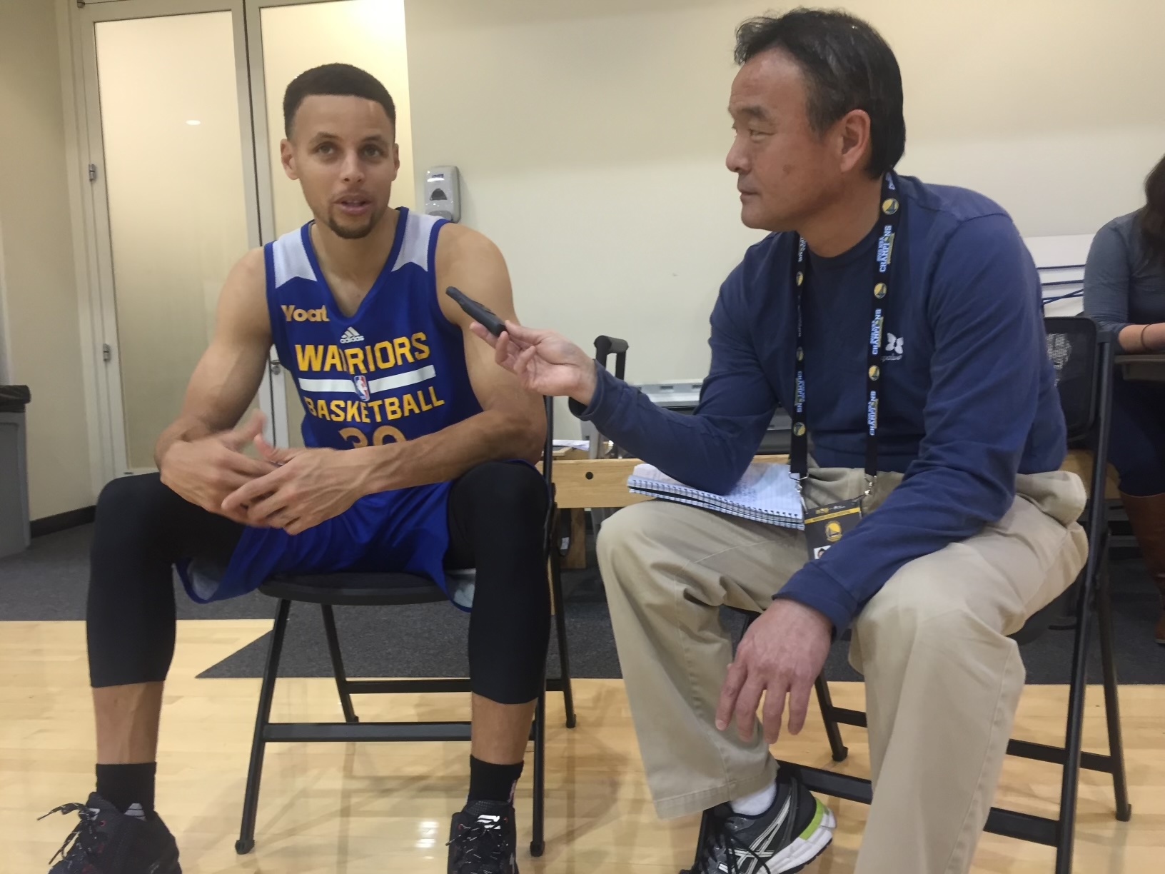 A basketball player in a blue Warriors jersey sits and speaks while a man in casual attire interviews him, holding a recorder and a notebook.