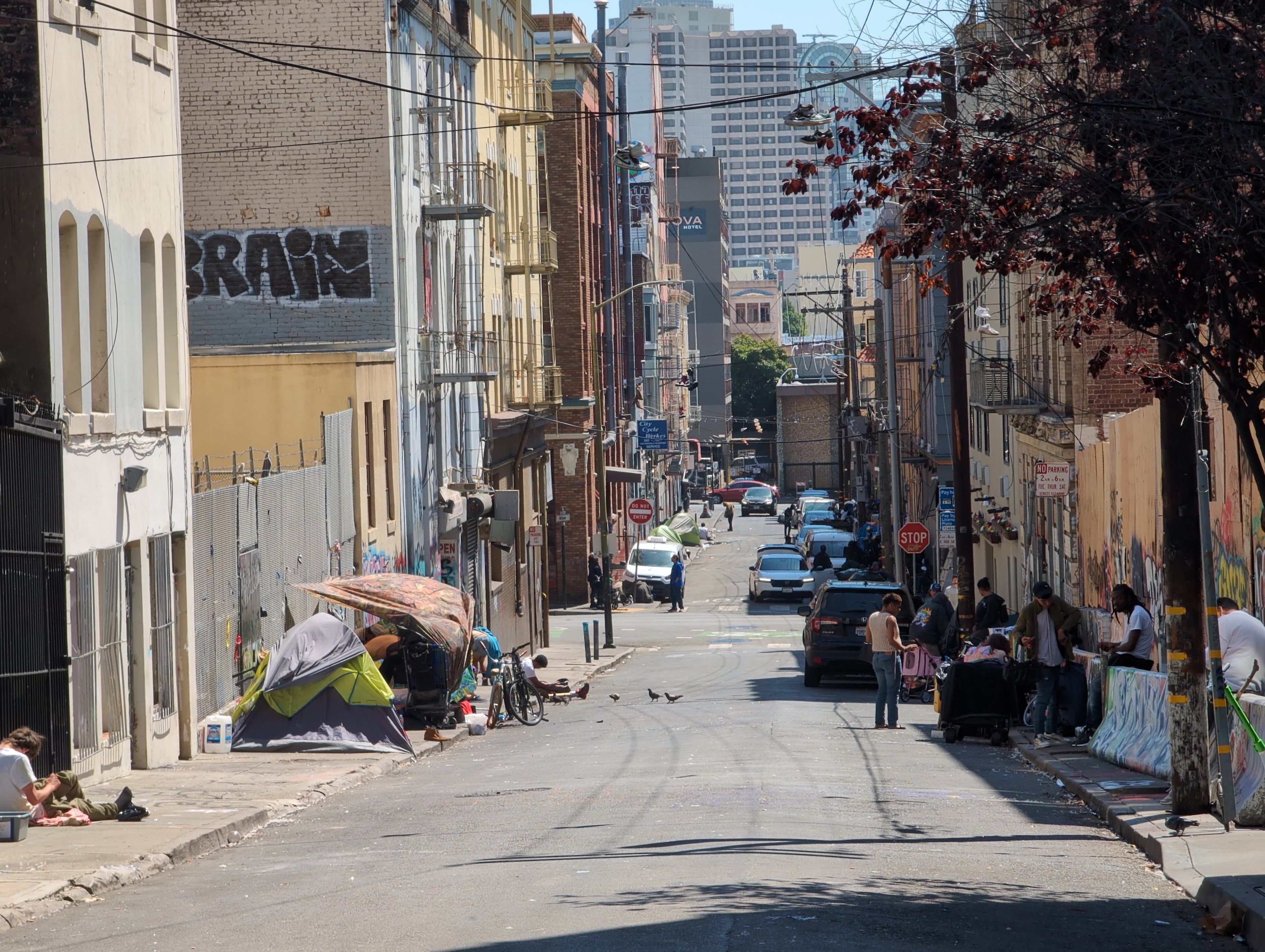 A city street with tents and people on the sidewalks. Buildings with graffiti line both sides, some damaged. Cars are parked, and people are interacting.