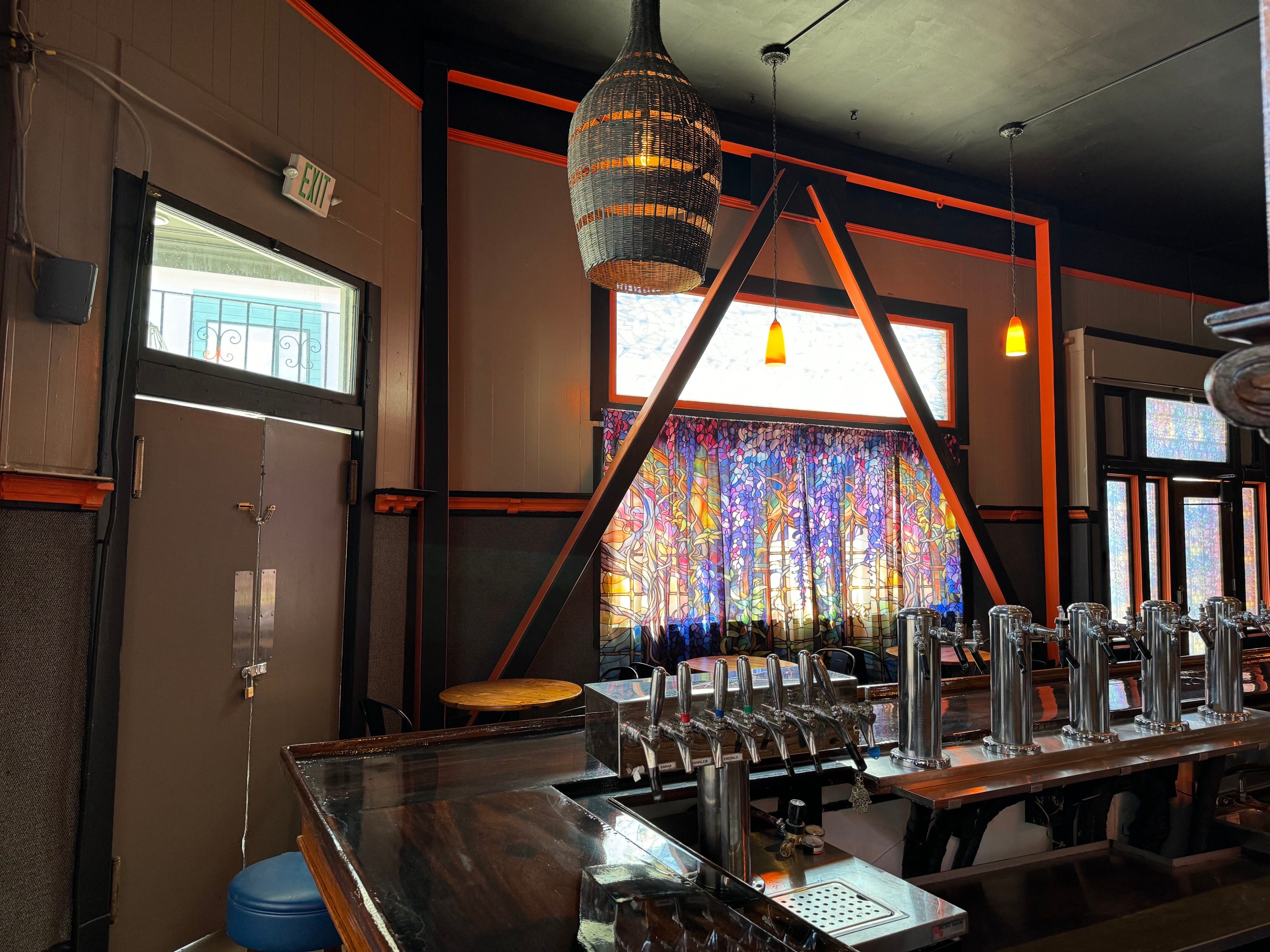 This image shows a bar with a wooden counter, multiple beer taps, colorful stained-glass window, a triangular wooden frame, a gray door, and hanging orange lights.