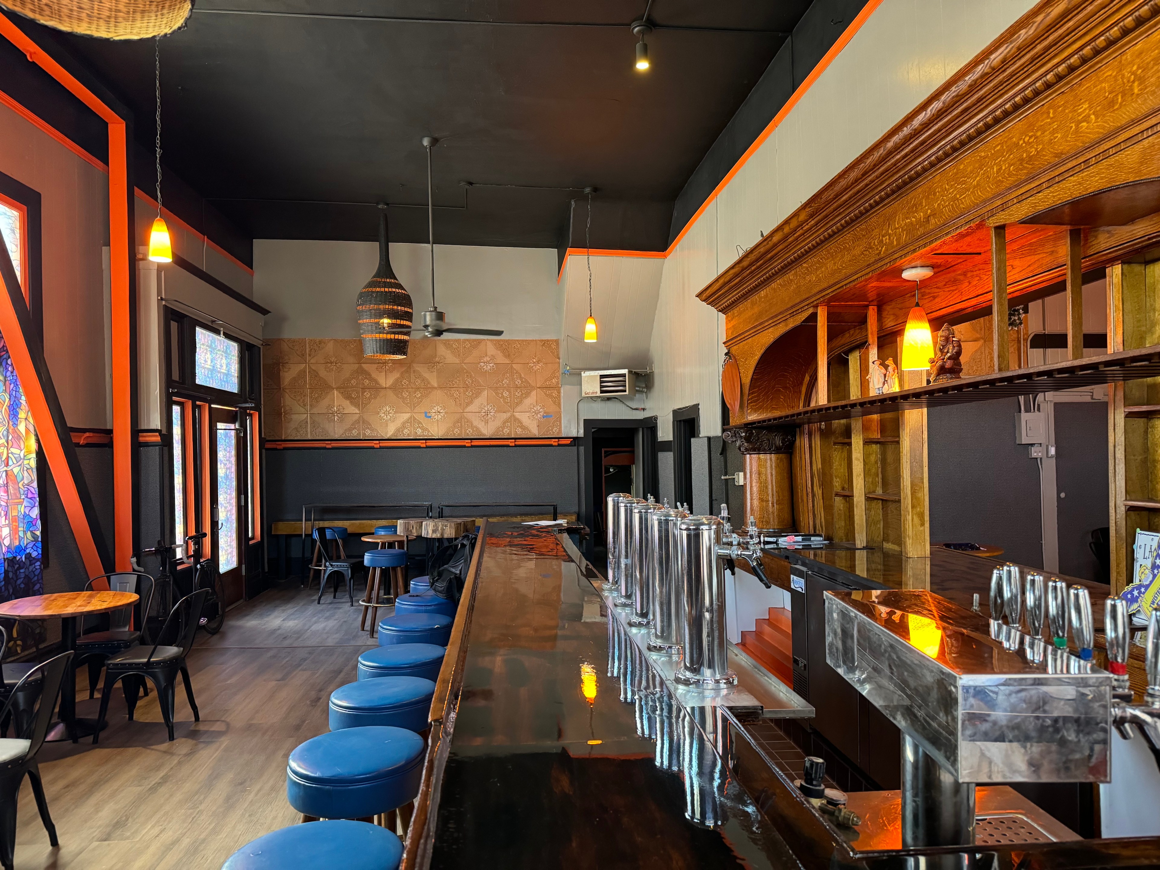 The image shows a bar with wooden decor, orange accents, pendant lights, a mirrored counter, and blue stools. Stained glass windows and mosaic tiled walls adorn the space.