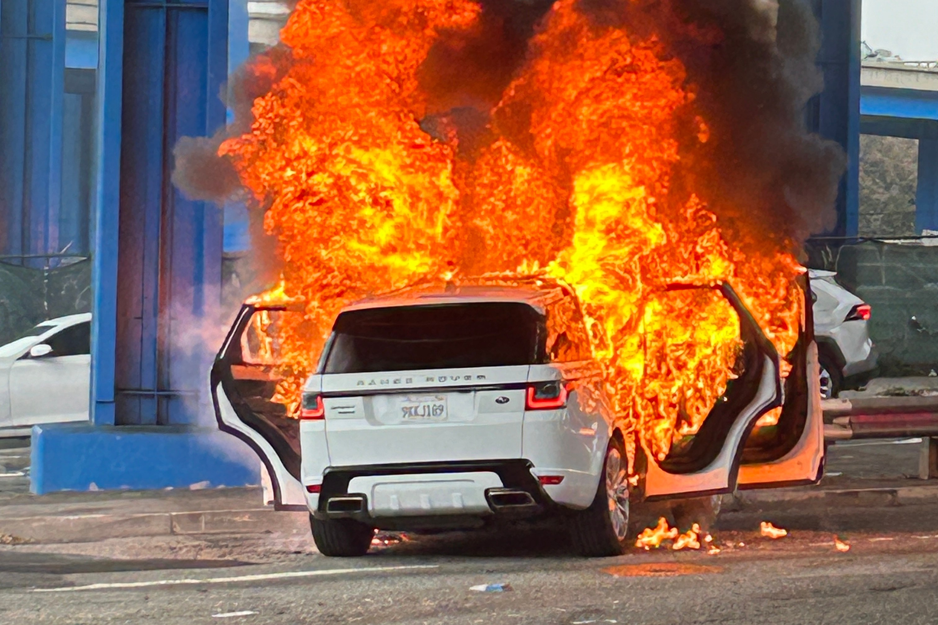 A white SUV is engulfed in flames with its doors open, emitting thick black smoke under a blue structural framework.