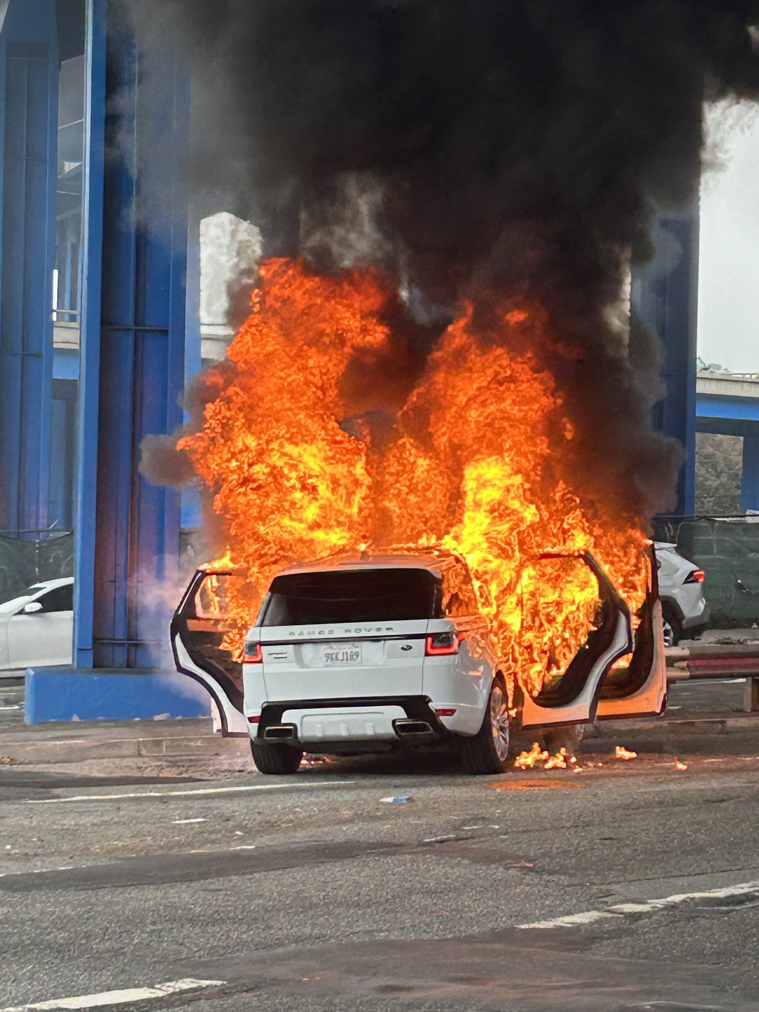 A white SUV is engulfed in flames with its doors open, emitting thick black smoke under a blue structural framework.