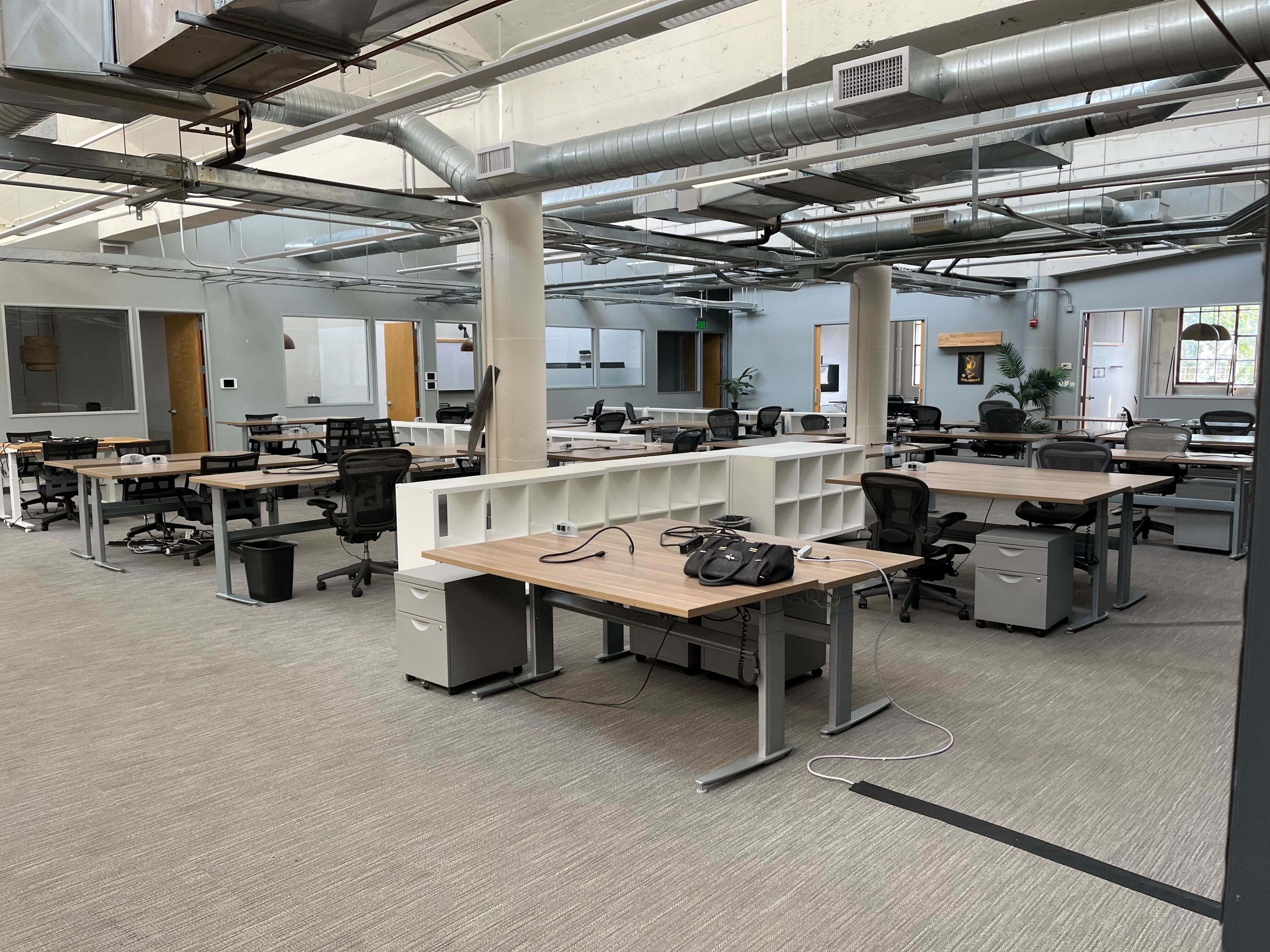 The image shows a spacious, open-plan office with various empty desks, chairs, overhead ducts, and ceiling pipes, creating an industrial look.