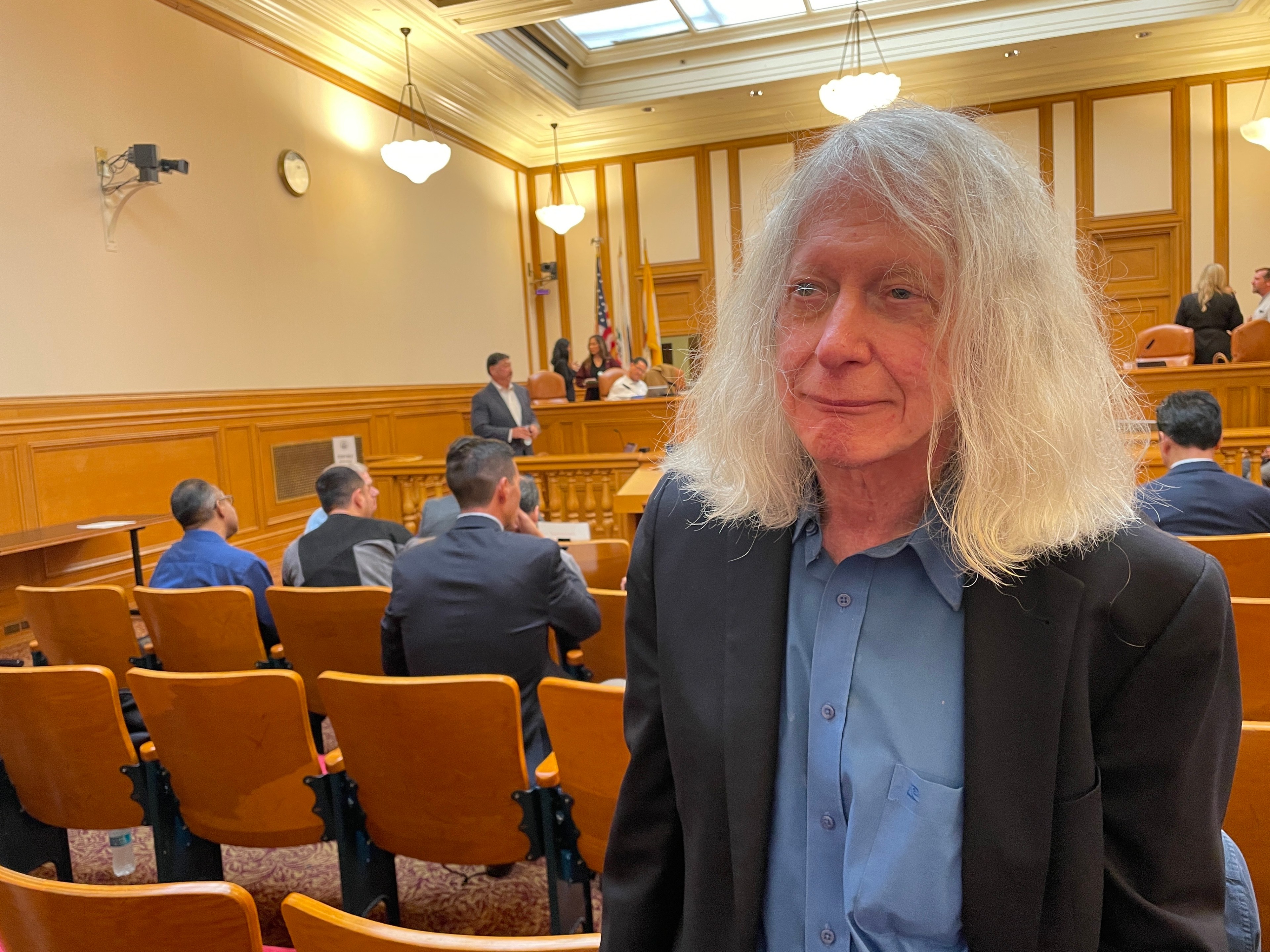 A man with long white hair, wearing a black jacket and blue shirt, stands in the foreground of a courtroom filled with people seated and someone speaking up front.