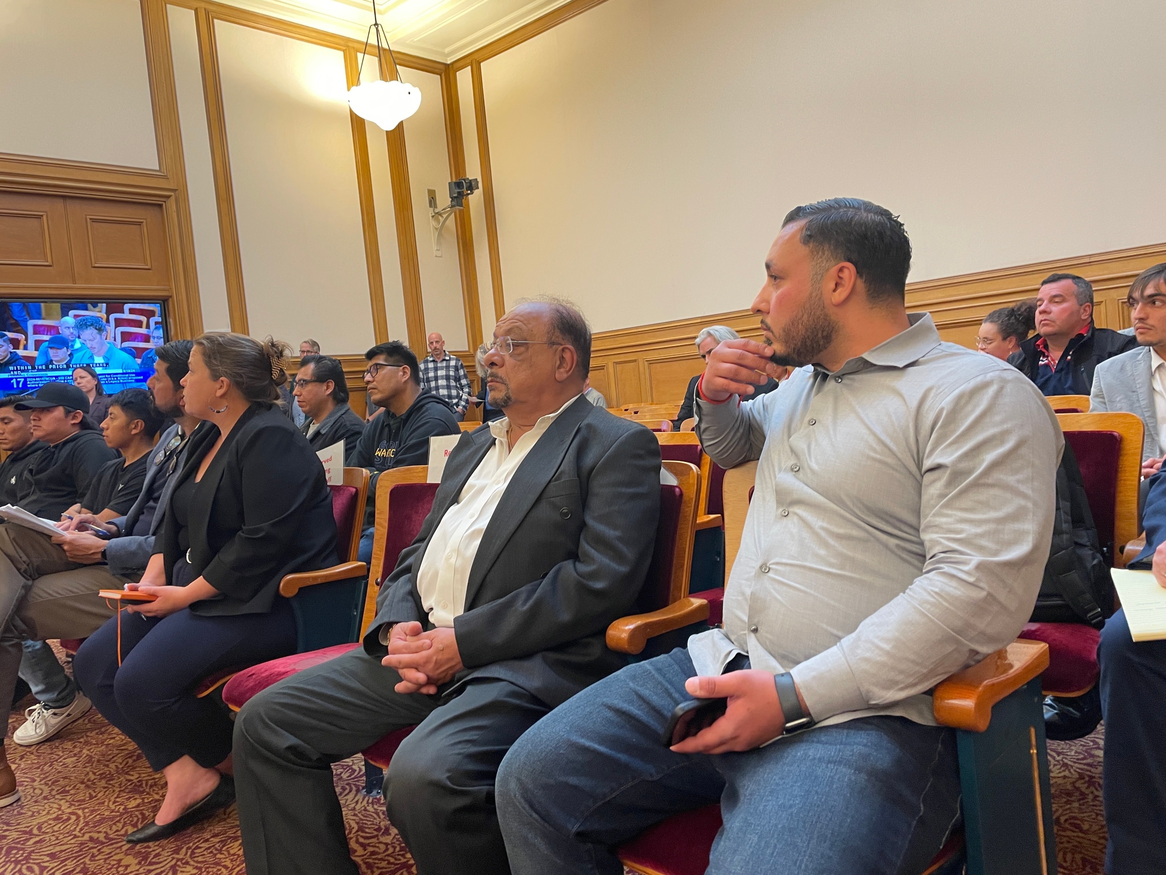 A group of people sit in a formal setting, such as a courtroom or meeting hall. They appear attentive, with some holding papers and notebooks while watching proceedings.