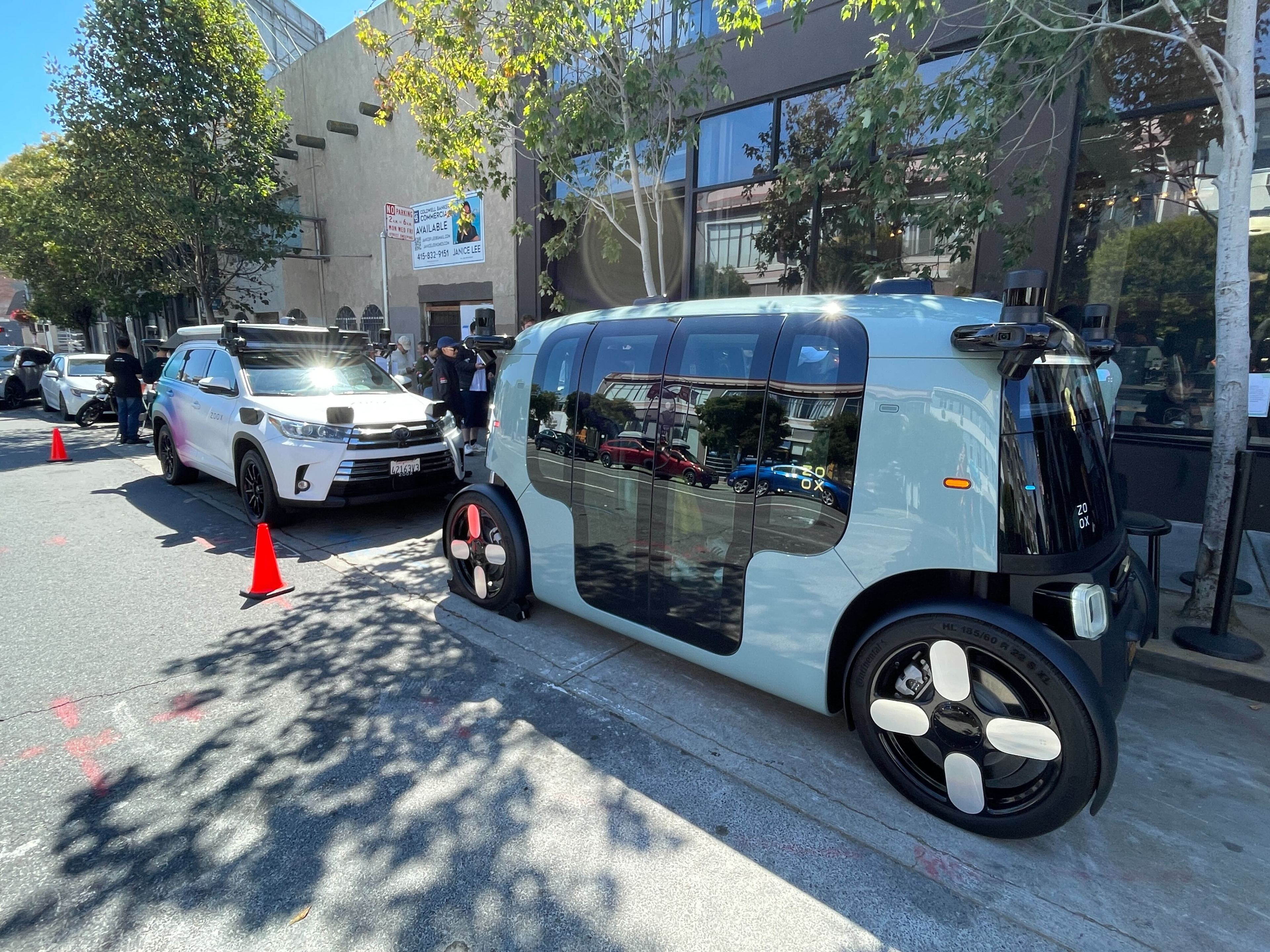 On a city street, there are two futuristic vehicles parked; one white SUV and one rounded, compact, self-driving pod car with several onlookers nearby and orange cones.