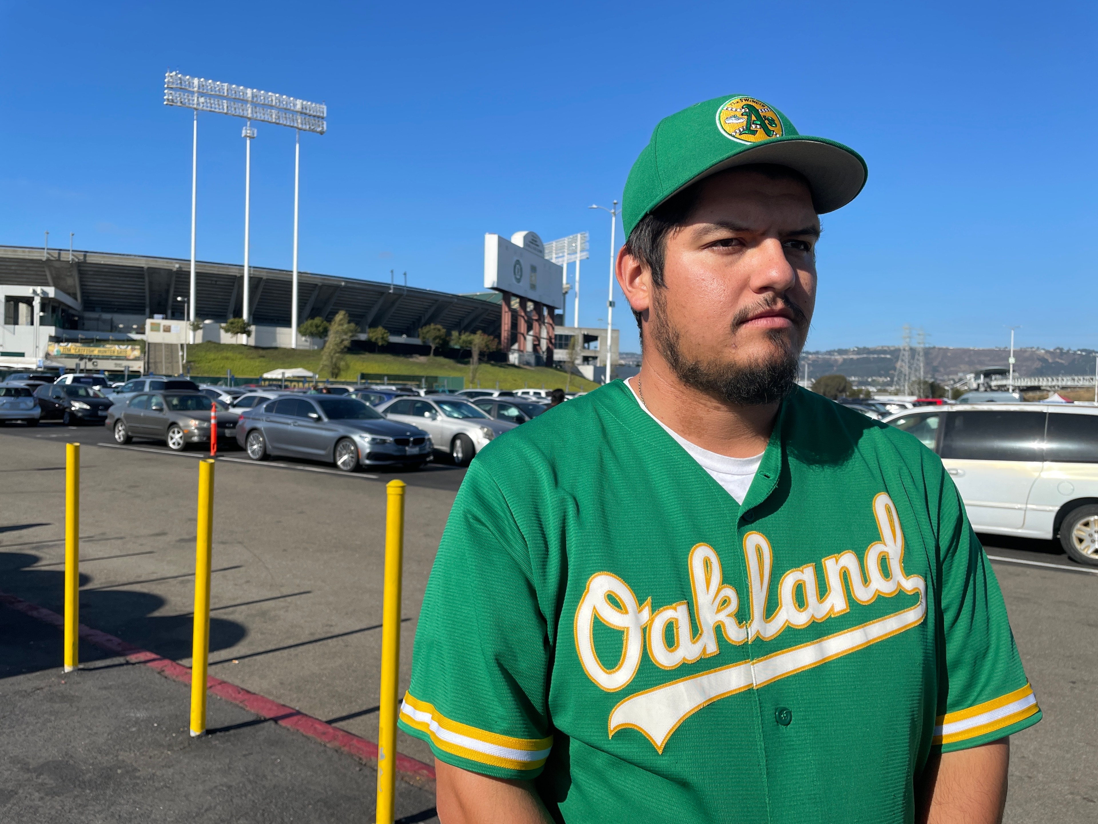 A man in a green baseball jersey and cap stands in a parking lot with a large stadium visible in the background under a clear blue sky.