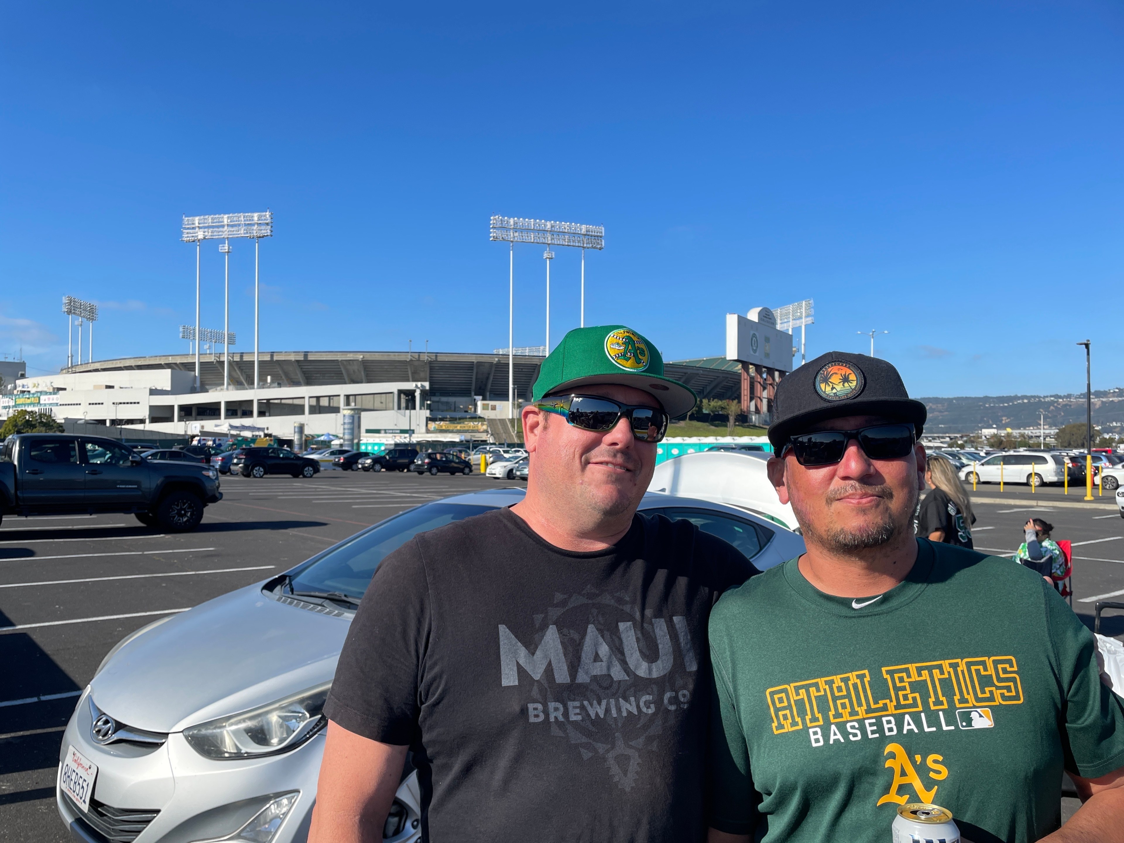 Two men in baseball hats stand in a parking lot with a stadium in the background. One wears an &quot;Athletics Baseball&quot; shirt, and they're smiling under a clear blue sky.