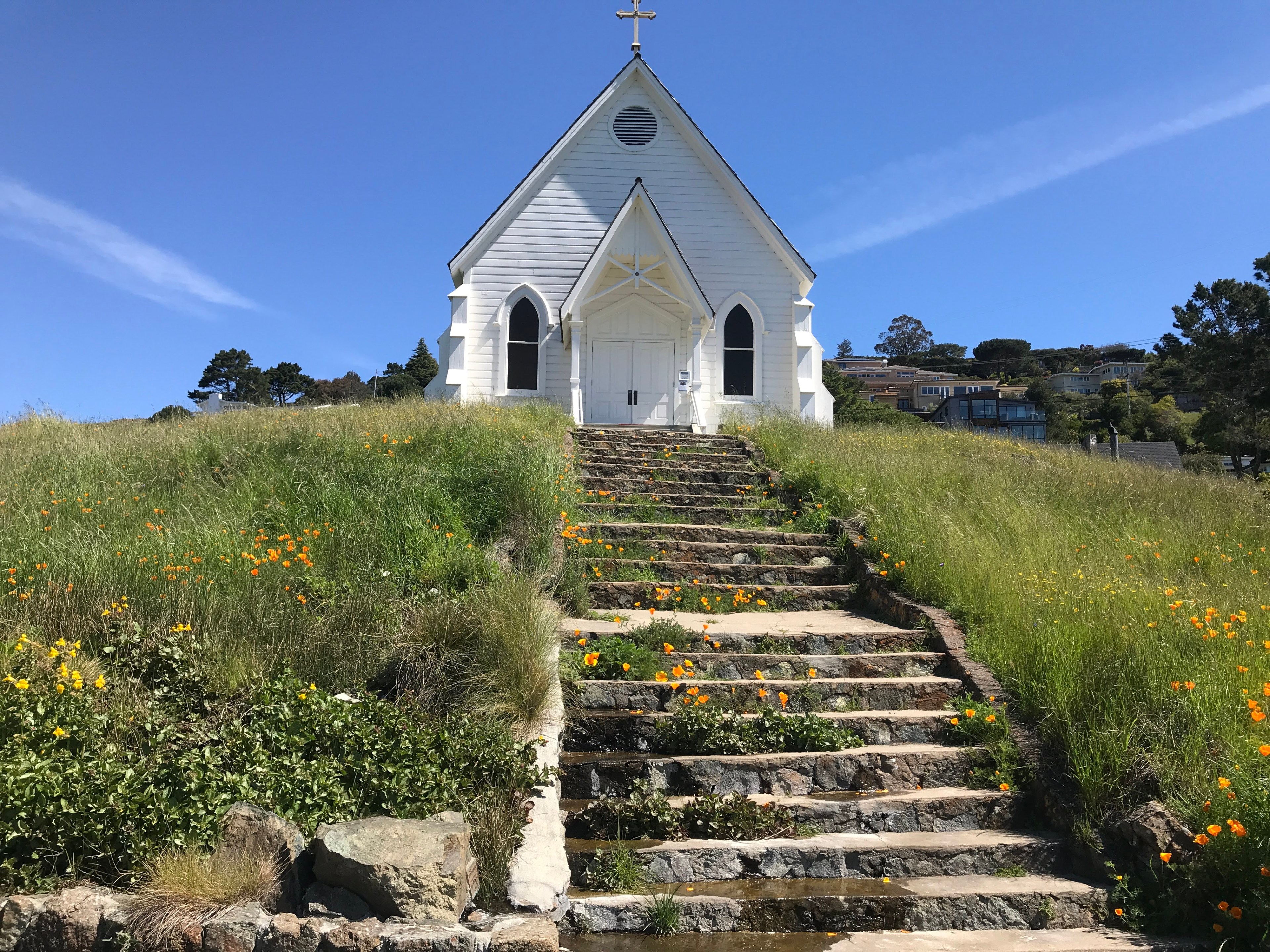 A quaint white chapel sits atop a hill, accessed by stone steps bordered with vibrant orange and yellow wildflowers, against a backdrop of clear blue sky and trees.