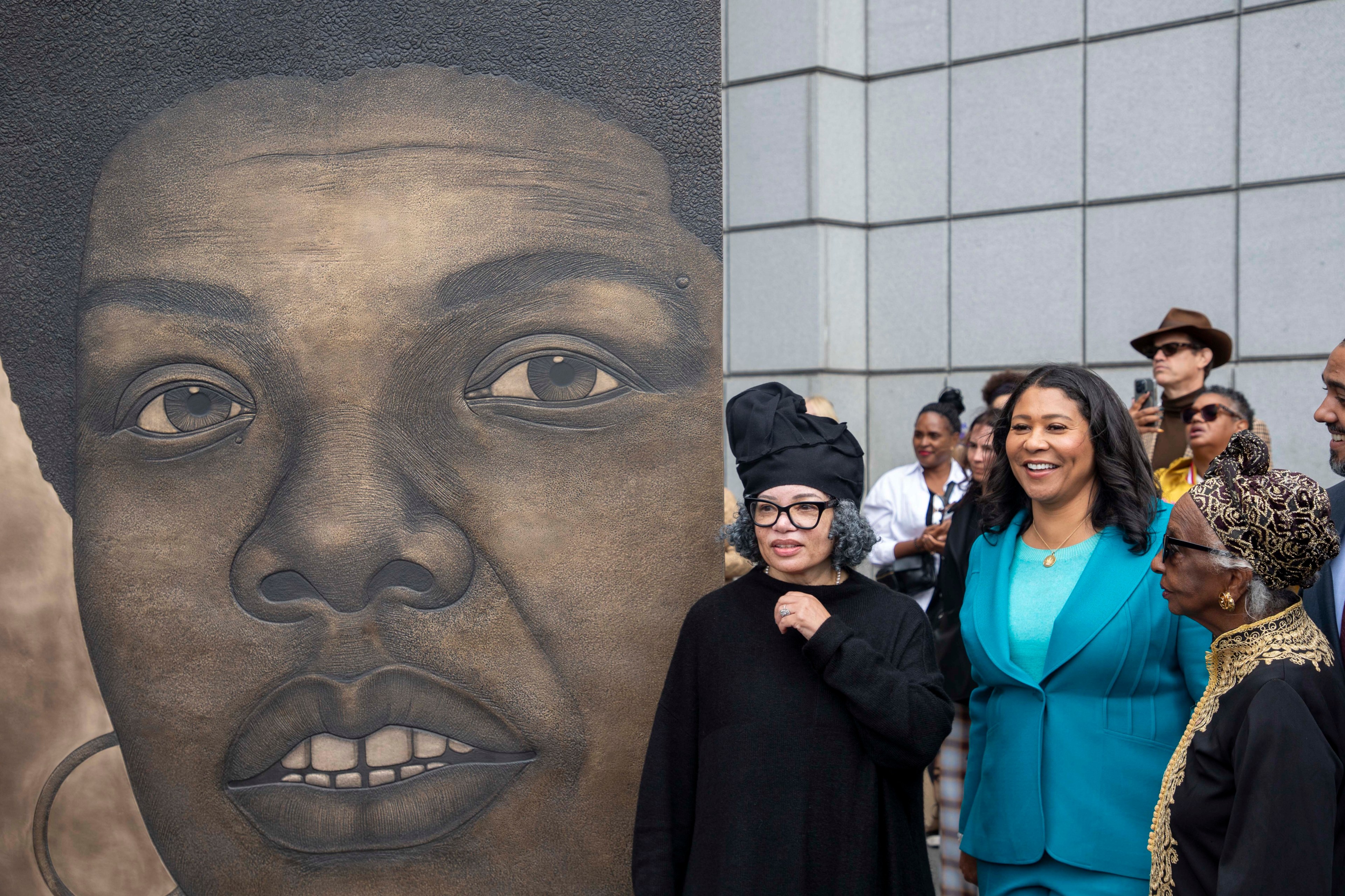 A group of people stand beside a large, detailed mural of a woman's face on a city sidewalk, with one woman in a bright teal outfit smiling.