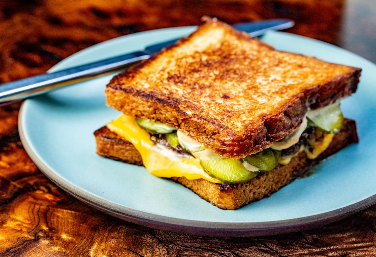 A toasted sandwich with cheese, pickles, and sauce on golden-brown bread sits on a light blue plate with a knife and fork beside it on a wooden table.