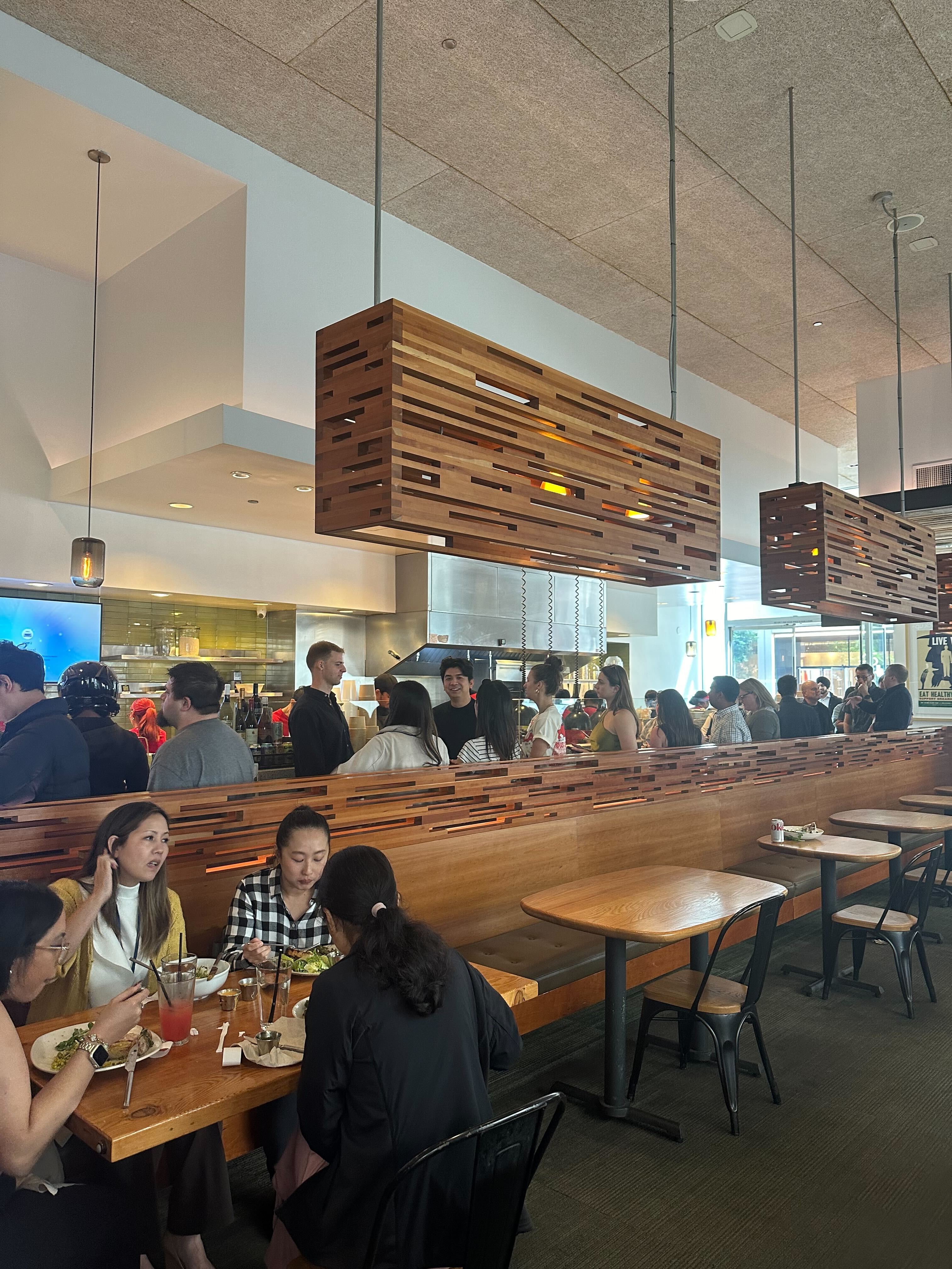 A bustling restaurant with wooden booths and modern lighting, where a line of diners stands at a counter, and a group of people are eating and conversing at a table.