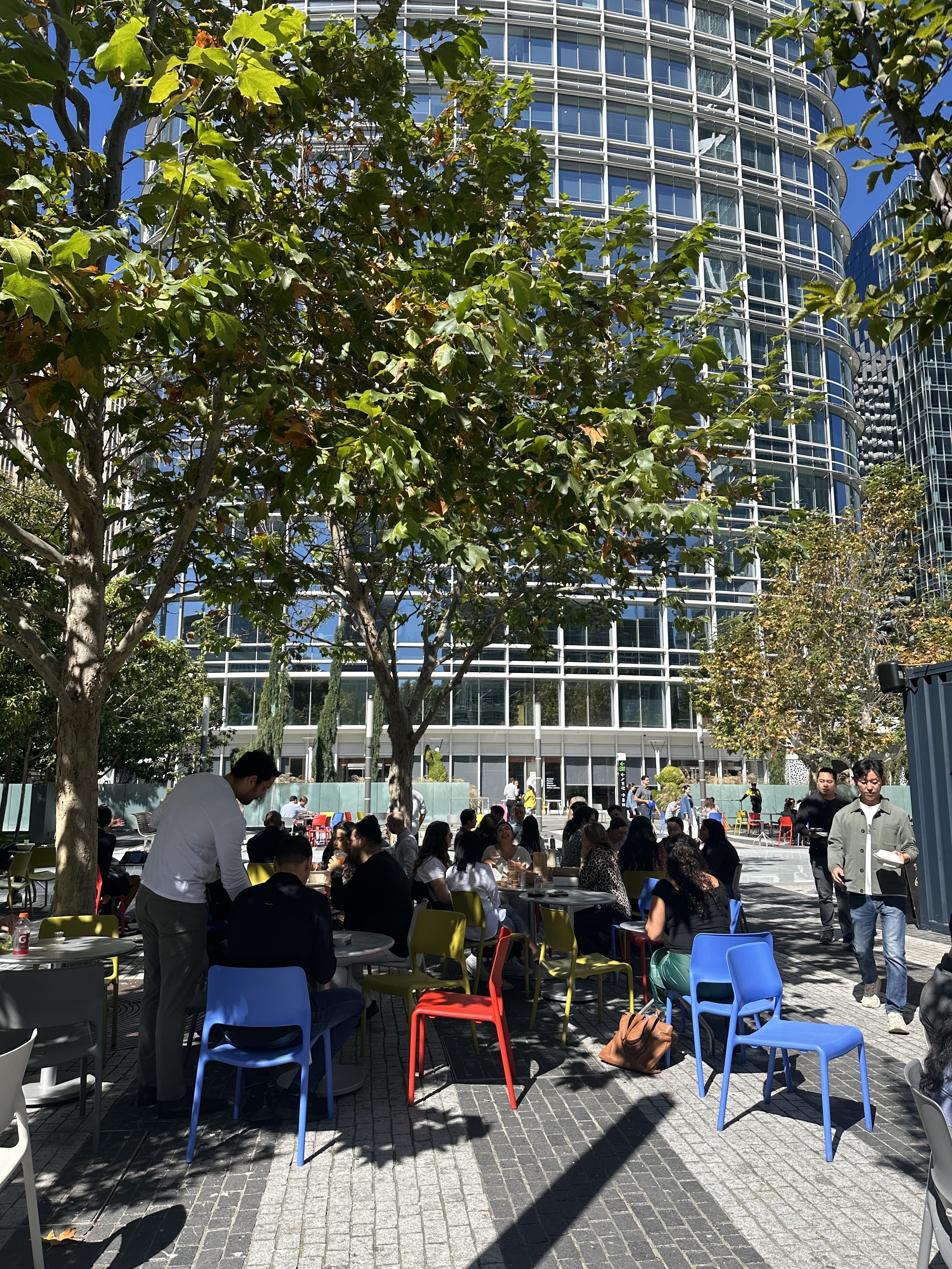 People are dining outdoors under leafy trees, seated on colorful chairs, with a modern building in the background.