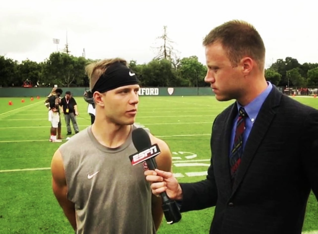 A man in a gray sleeveless shirt and headband is being interviewed on a football field by another man in a suit holding an ESPN microphone.