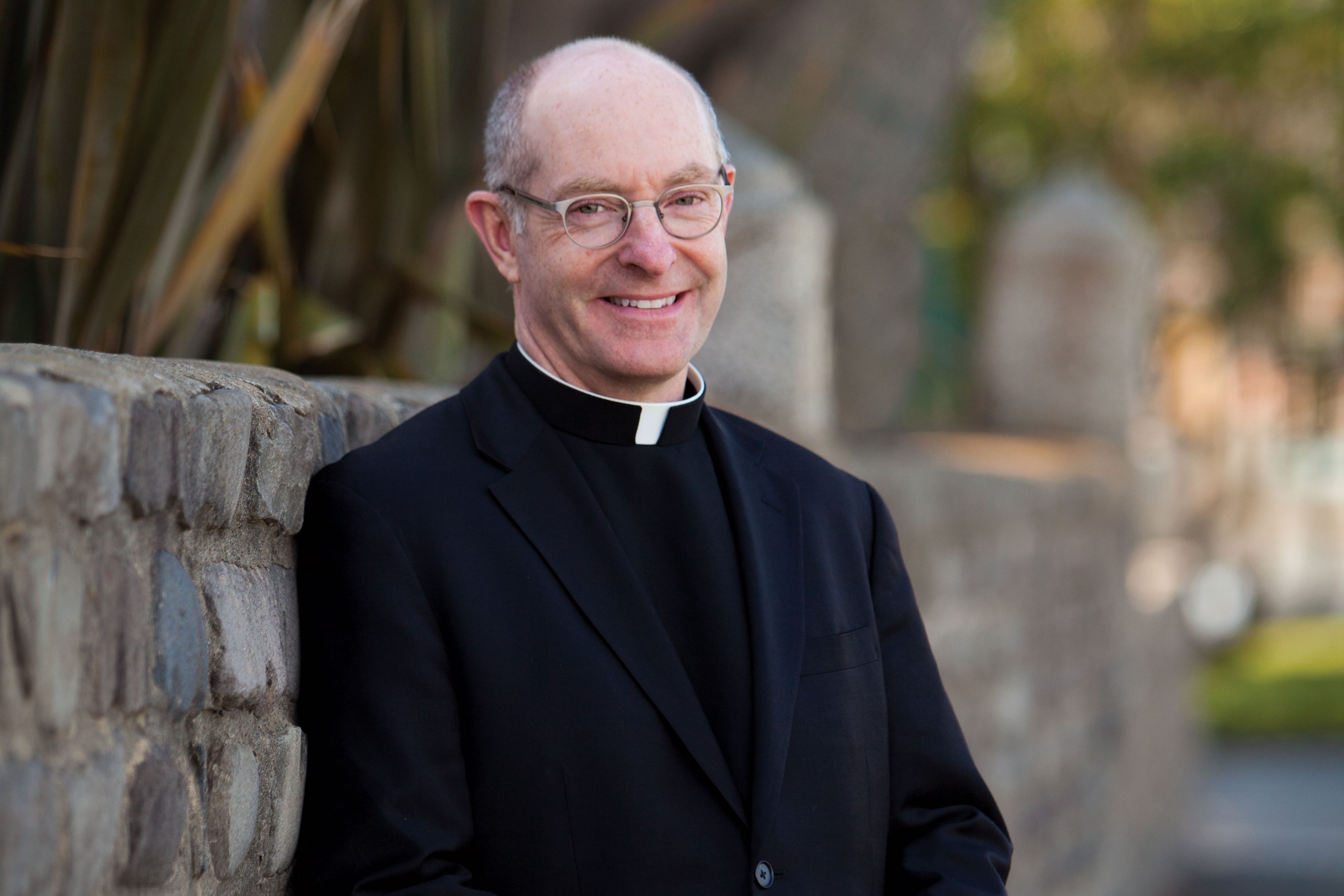 A smiling man wearing glasses and clerical attire with a white collar stands against a stone wall with plants in the background.