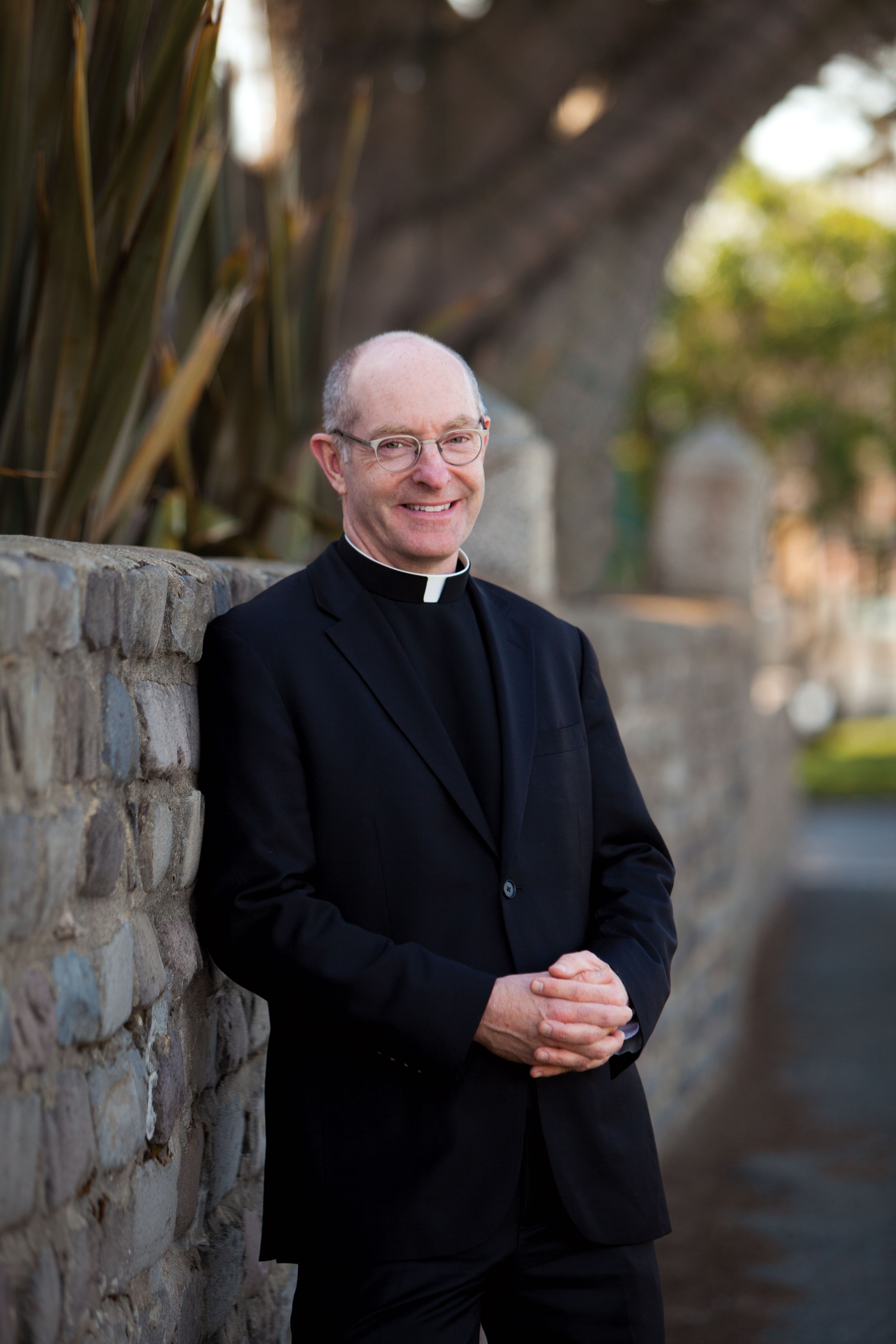 A smiling man wearing glasses and clerical attire with a white collar stands against a stone wall with plants in the background.