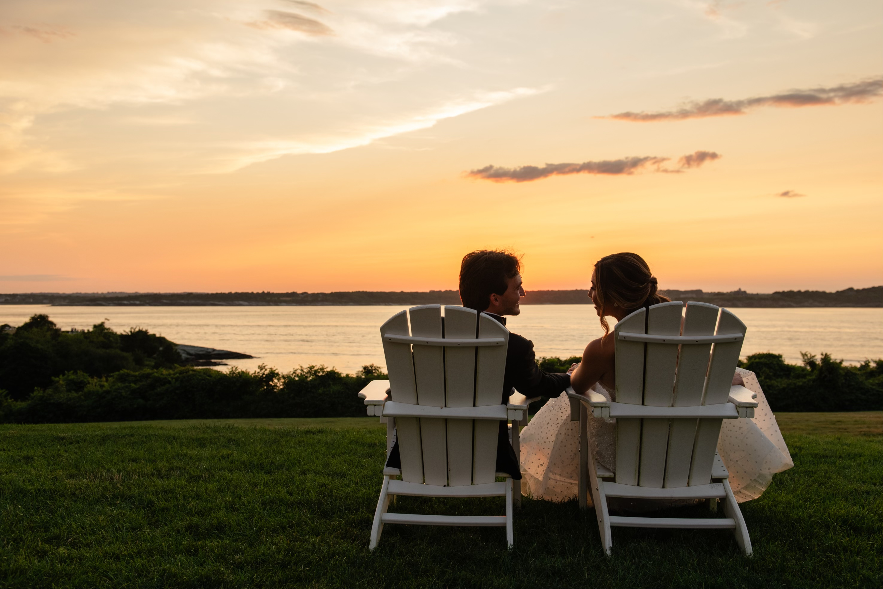 A couple sits in white chairs on a grassy hill, holding hands and gazing at a serene sunset over a calm body of water.