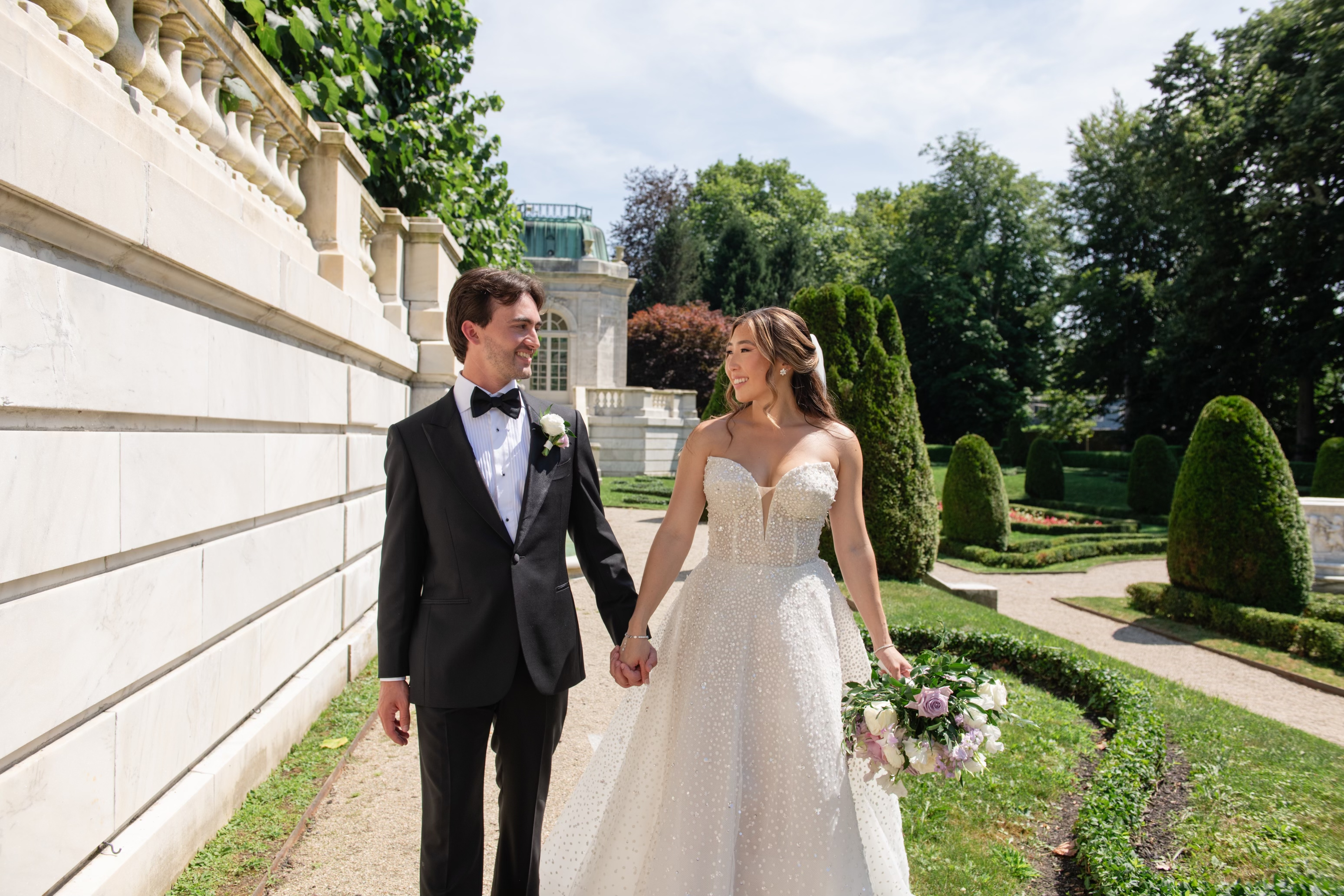 A bride and groom hold hands, smiling at each other, walking in a formal garden. The bride wears a white gown and holds a bouquet; the groom is in a black tuxedo.