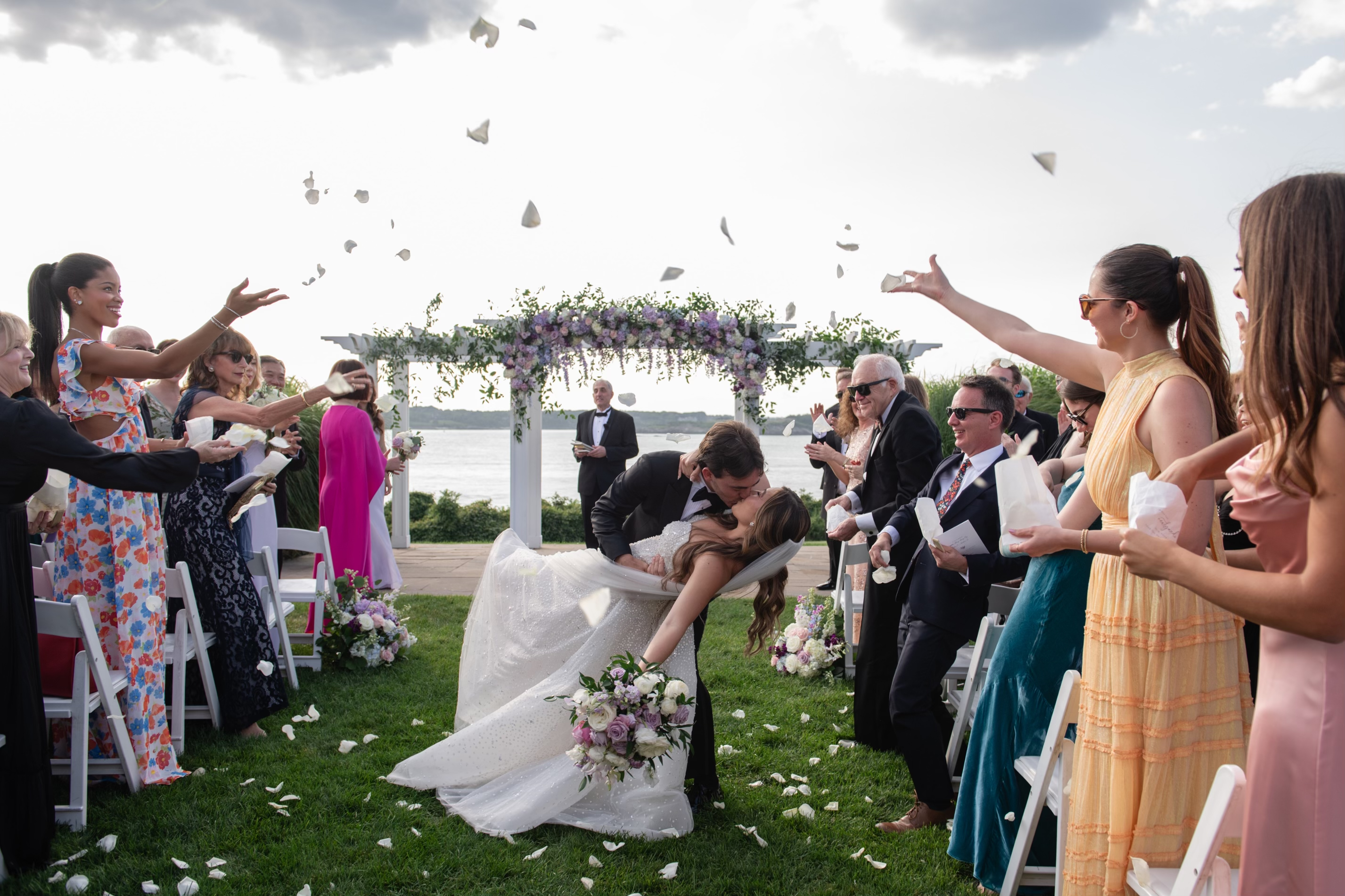 A couple kisses under a floral arch while guests toss flower petals. The event is outdoors by a waterfront, with attendees dressed in elegant attire.