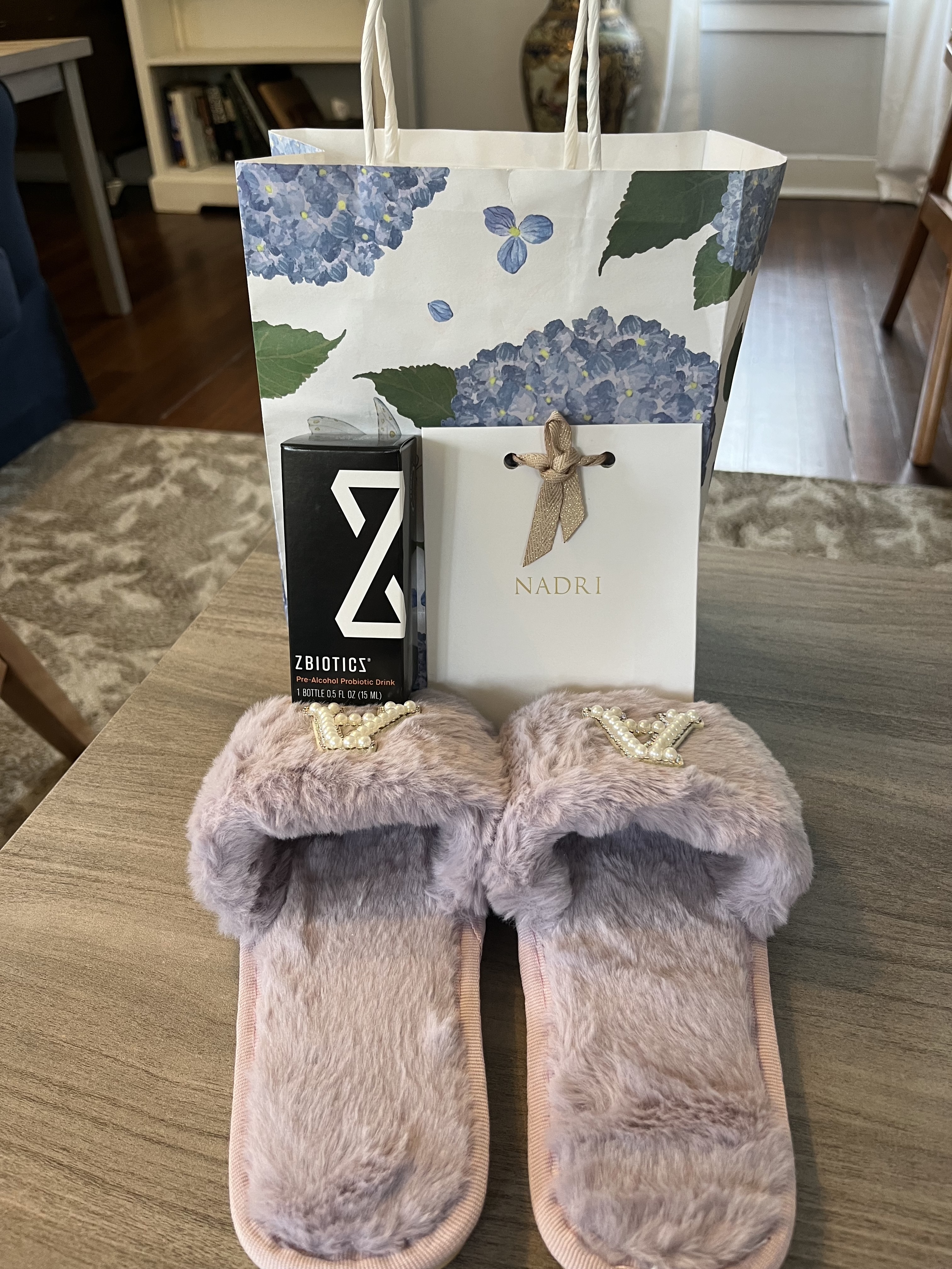 A pair of plush, lavender slippers, a black Zbiotics box, and a Nadri gift bag with a floral design and gold bow sit on a table in a cozy, furnished room.