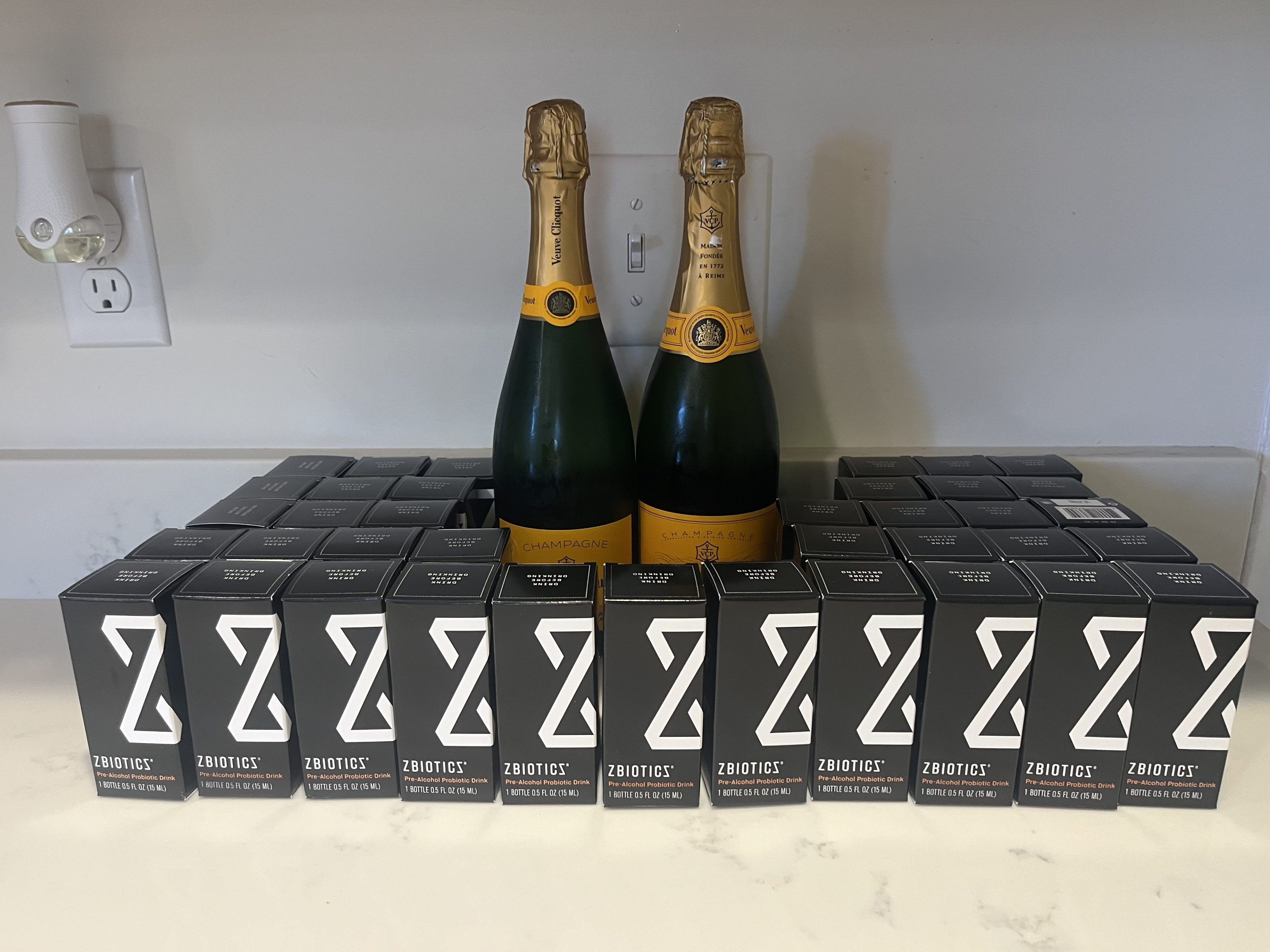 There are two bottles of Veuve Clicquot Champagne surrounded by numerous black boxes labeled &quot;Zbiotics&quot; on a marble counter, beneath a wall outlet.