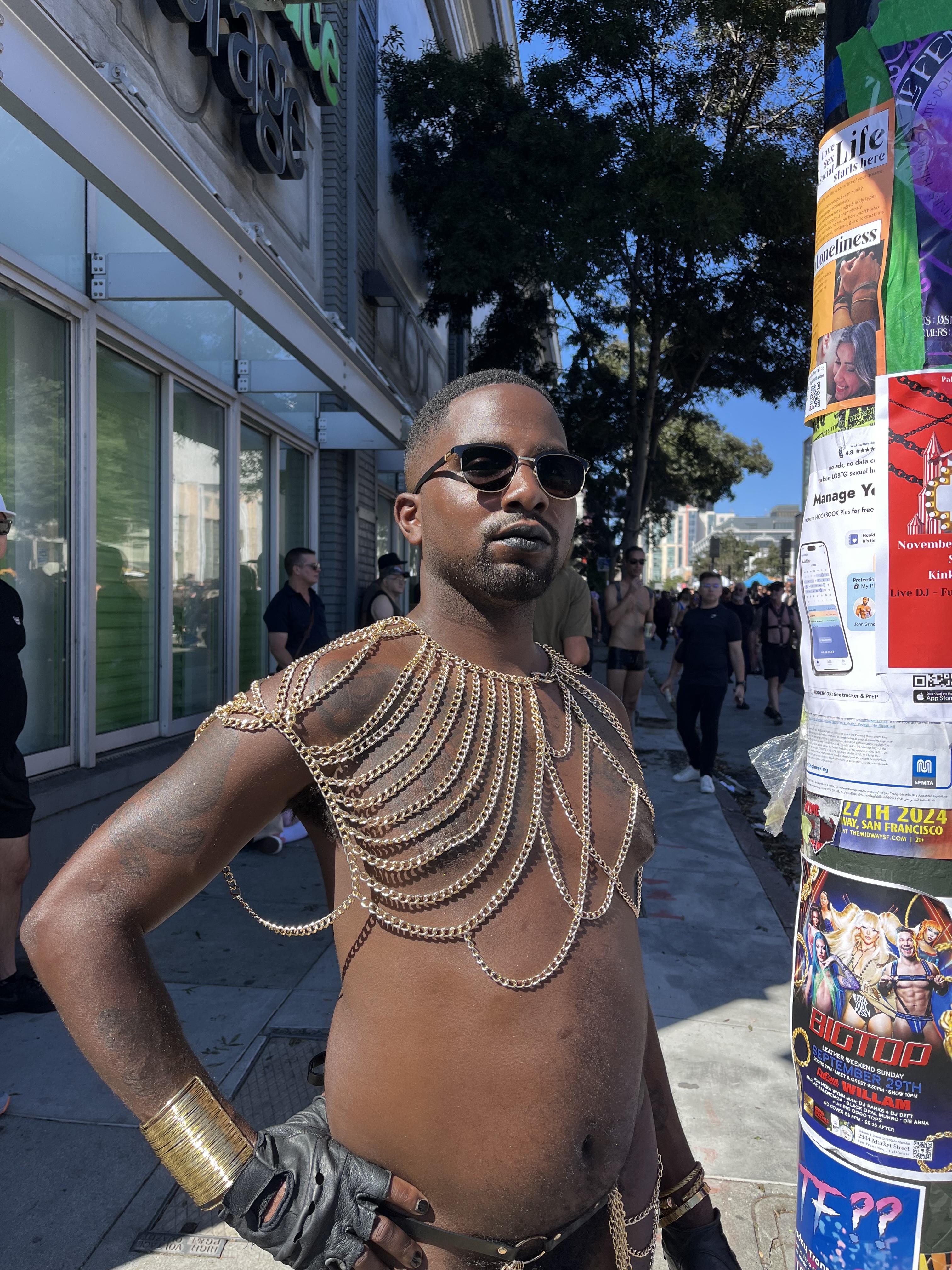 A confident individual in sunglasses and a gold chain harness poses on a street with various posters on a utility pole and people in the background.