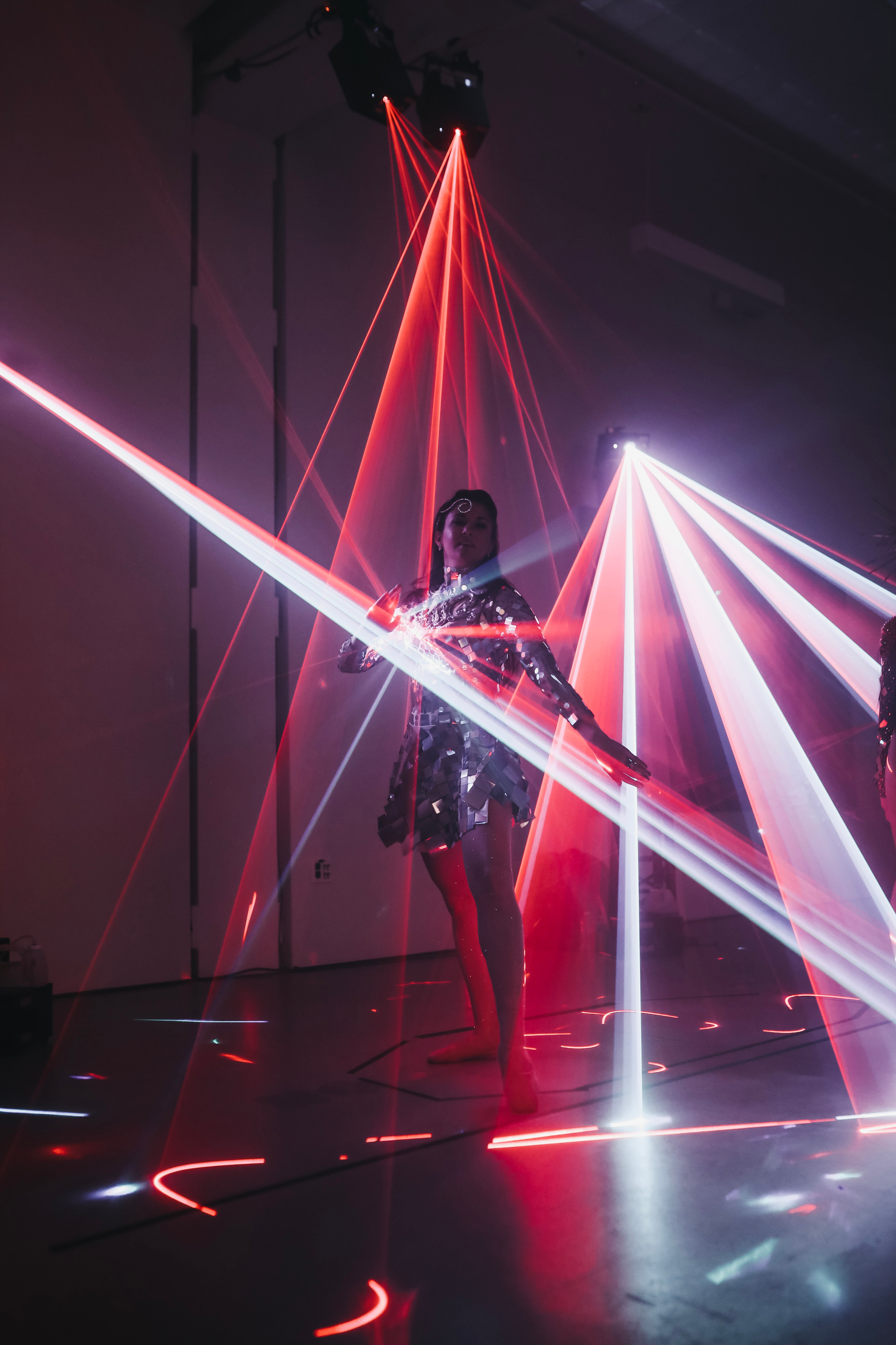 A person in a metallic outfit is standing among vibrant red and white laser beams, creating a visually striking and futuristic light display in a dimly-lit room.