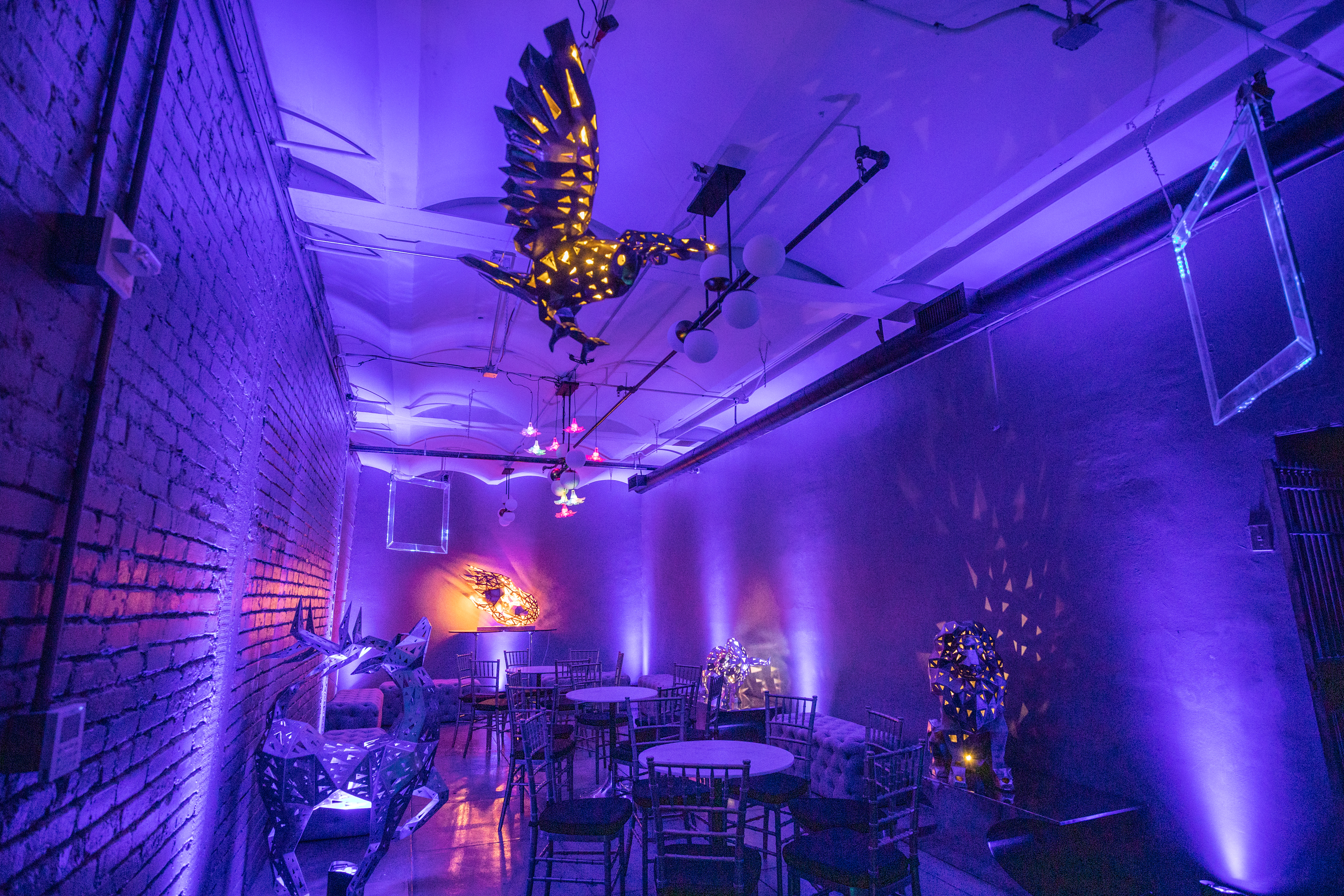 The room features purple-lit walls, modern art sculptures, round tables with chairs, a brick wall, and a ceiling adorned with an illuminated bird figure.