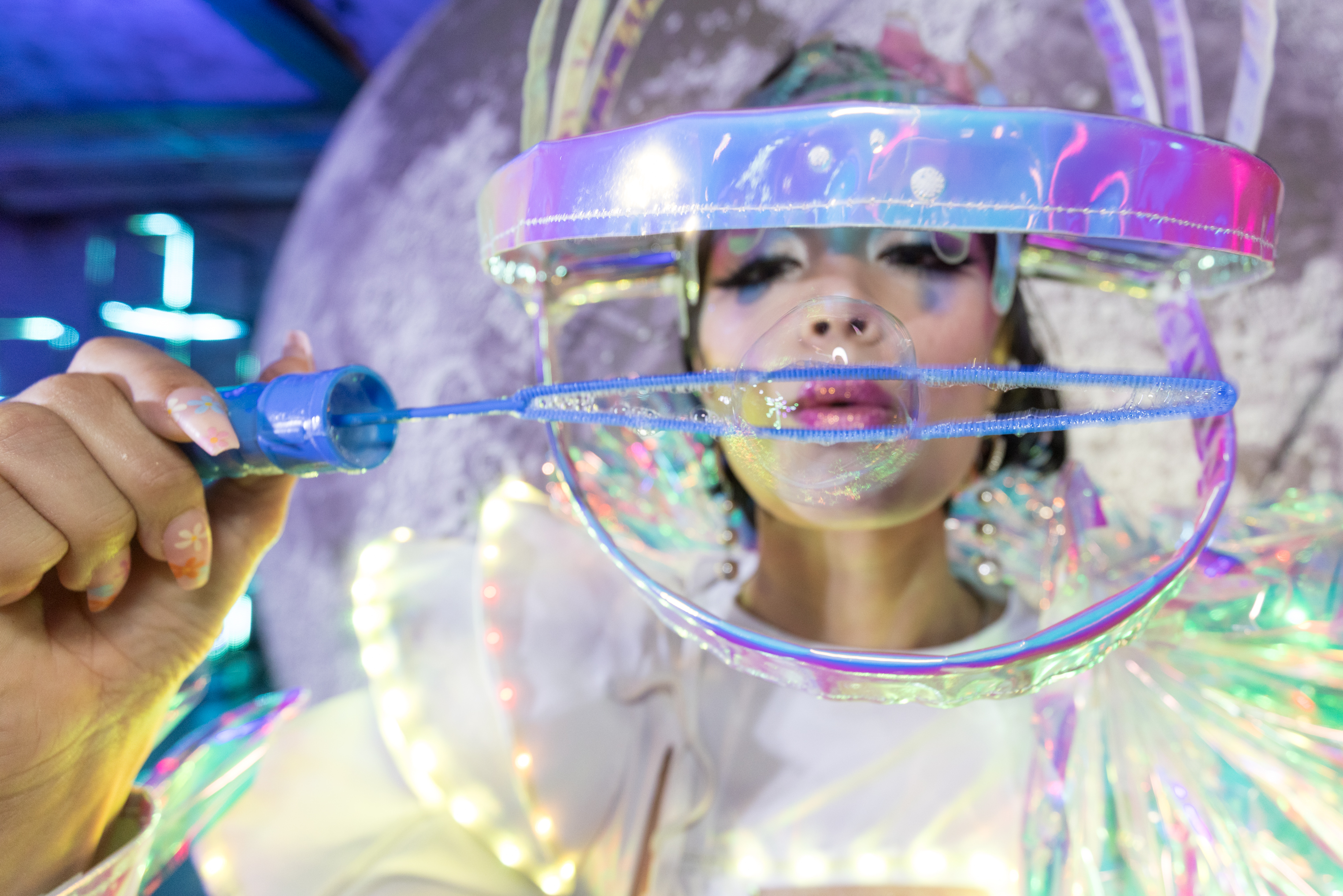 A person in futuristic, rainbow-themed attire blows a bubble using a blue bubble wand, with shiny makeup and iridescent light effects surrounding them.