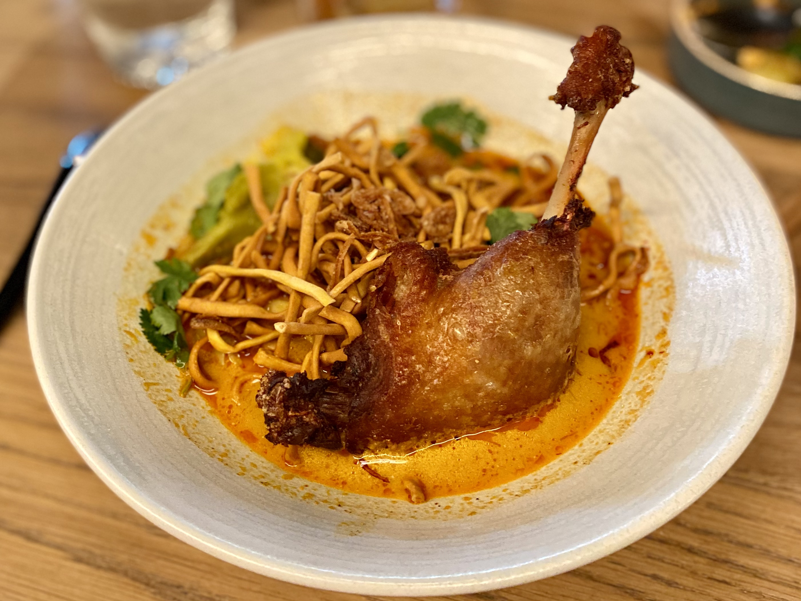 The image shows a bowl of flavorful curry with a crispy roast chicken leg, garnished with fresh herbs and crunchy fried noodles on top.