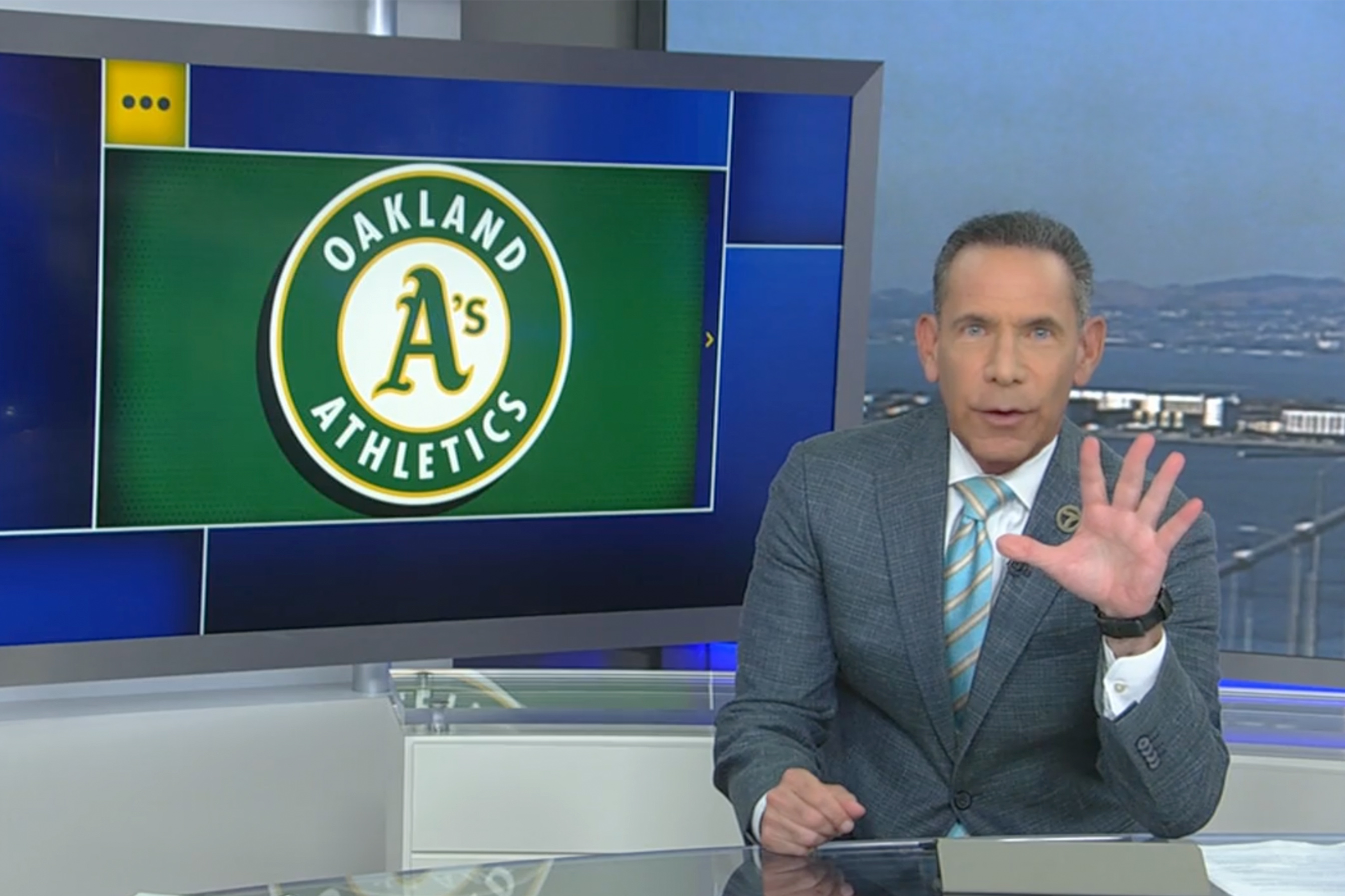 A man in a suit and tie is presenting news in a studio with a screen displaying the Oakland Athletics logo behind him.